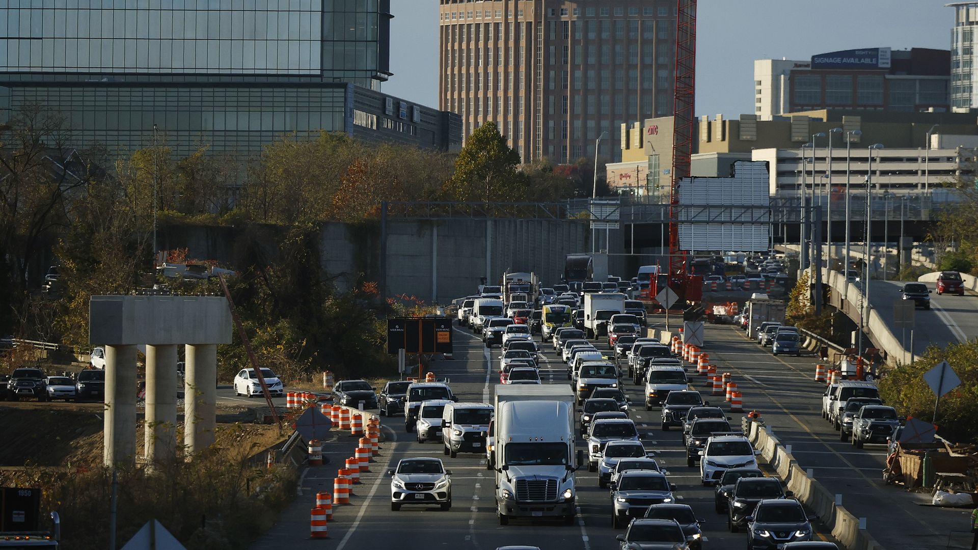Busy city highway with multiple lanes of traffic and orange construction cones, flanked by a glass office building on the left and a tan tower with arched tops; a red crane rises in the background.