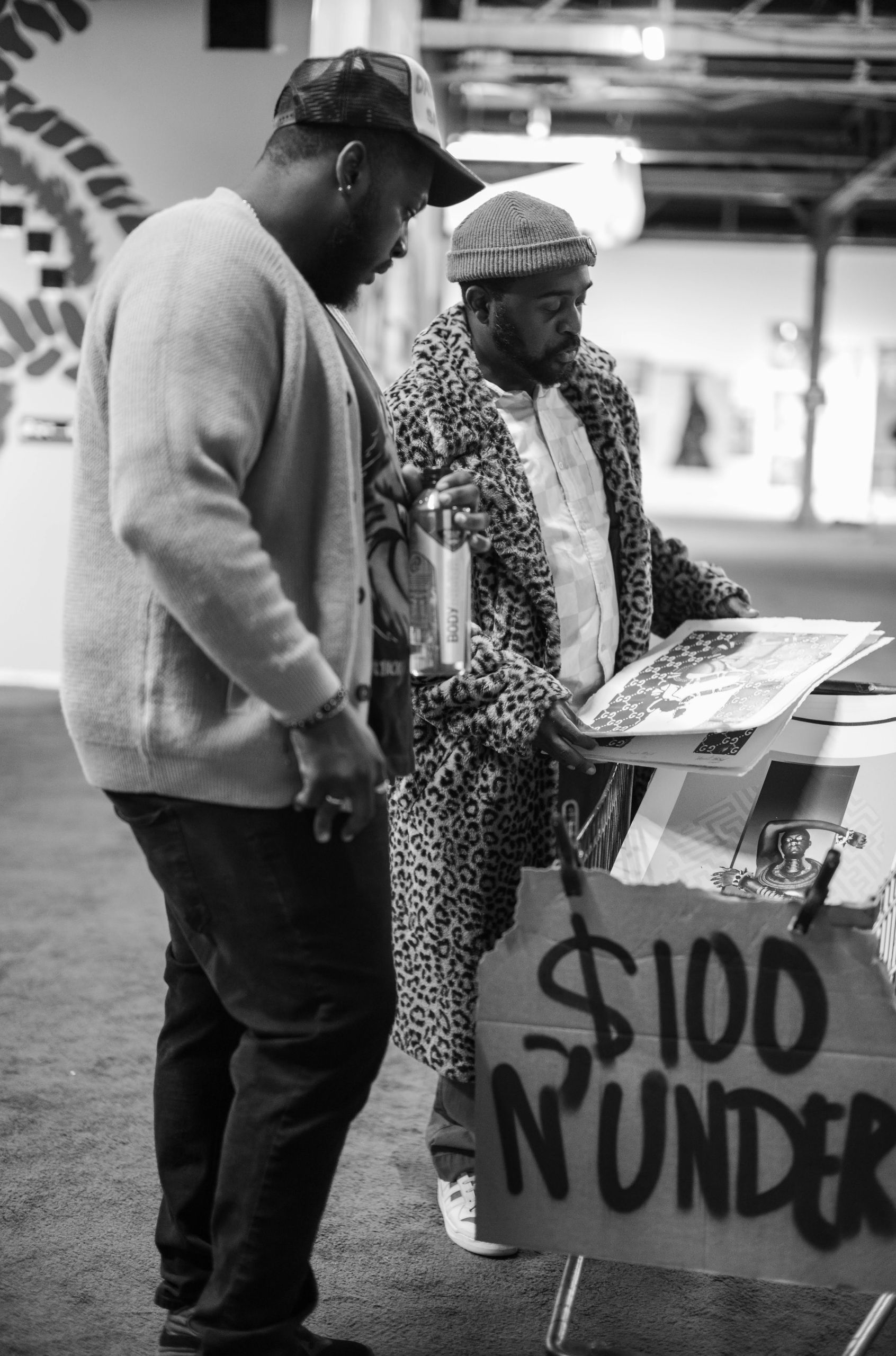 Two men examining artwork at a market or gallery; one wears a leopard print coat and beanie, the other a cardigan and cap holding a bottle. A sign nearby reads "$100 N' UNDER."