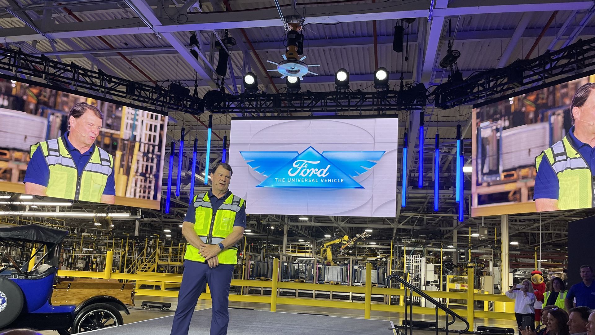 Man in a yellow safety vest stands on stage in a factory setting with a blue and white Ford logo displayed on a screen behind him and an audience watching in front.