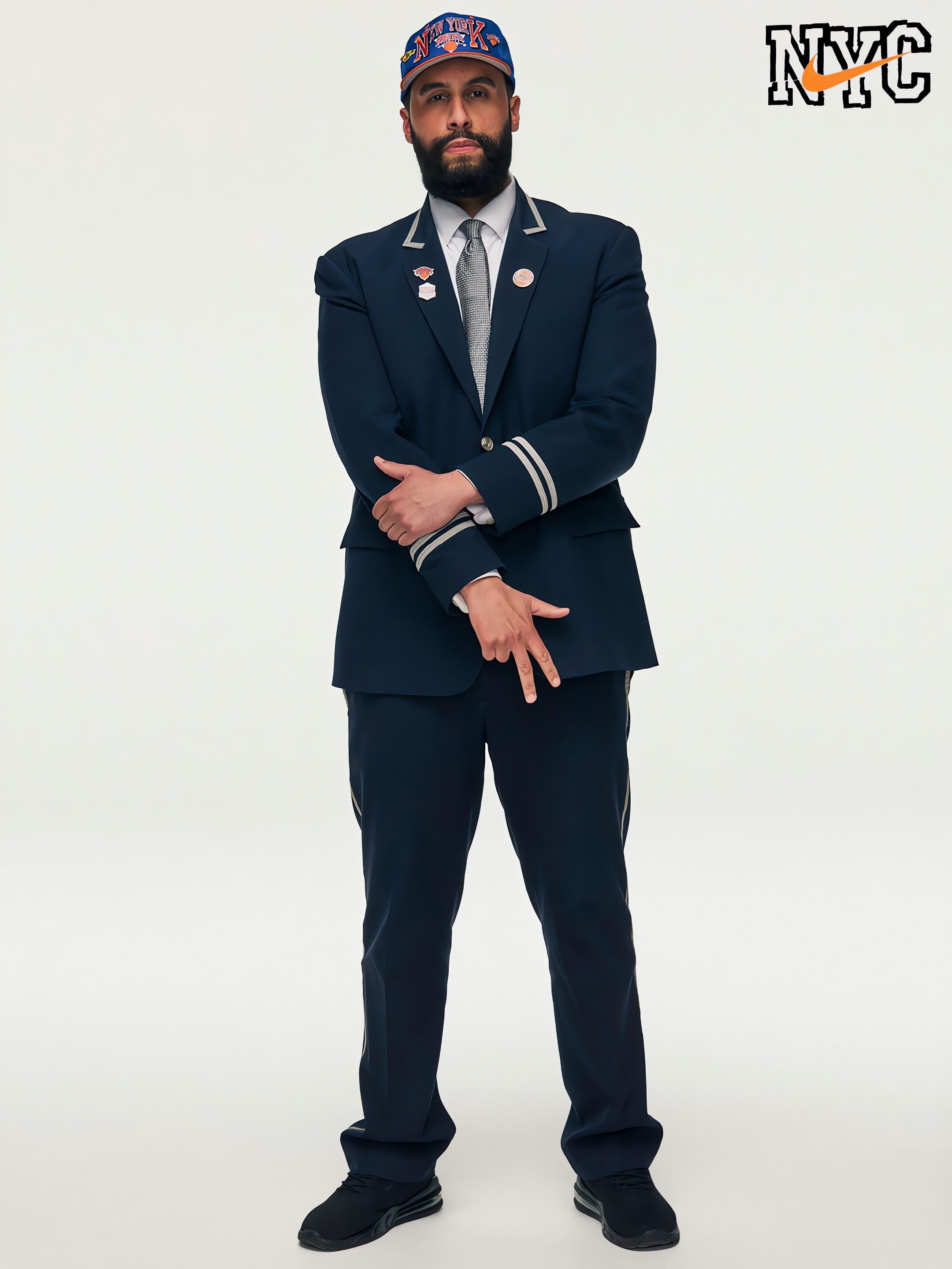 A man with a beard in a navy suit, white shirt, and gray tie, wearing a blue New York Knicks cap. He crosses his arms, lapel pins visible, against a plain white backdrop, with an NYC logo in the top right.
