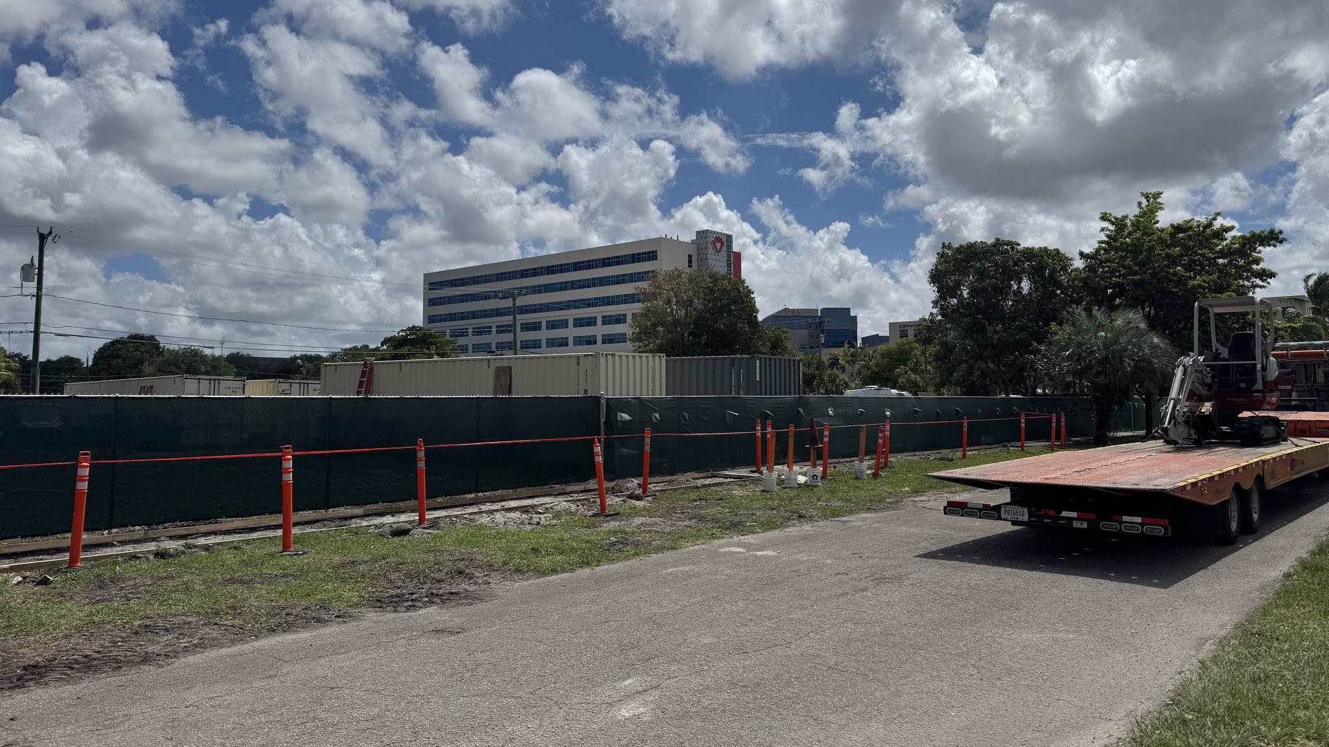 Construction site along a road with orange safety cones and a green barrier. In the background a white office building with a red heart logo, trees, and a blue sky with fluffy clouds.