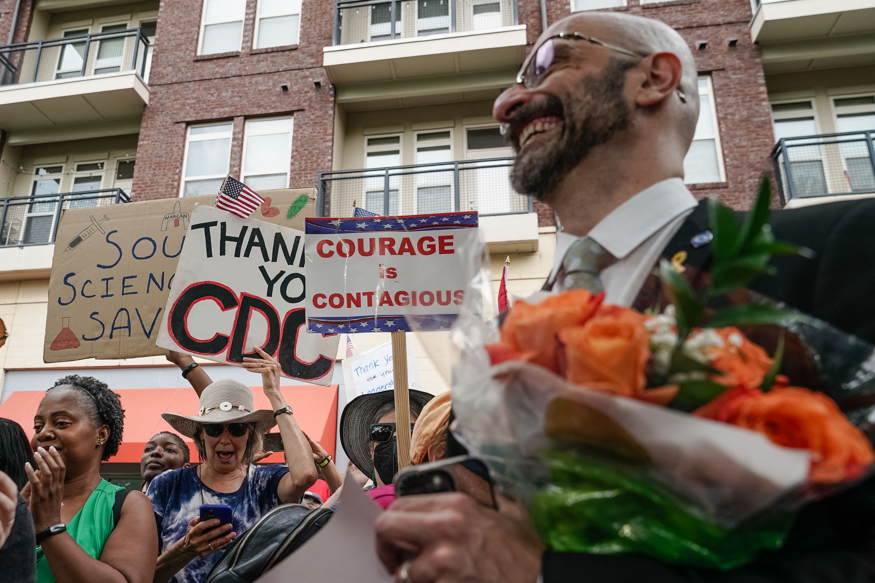 People at an outdoor event holding signs like "Thank You CDC," "COURAGE is CONTAGIOUS," and "Science Saves" while a man in a suit holds a bouquet of orange roses, smiling.