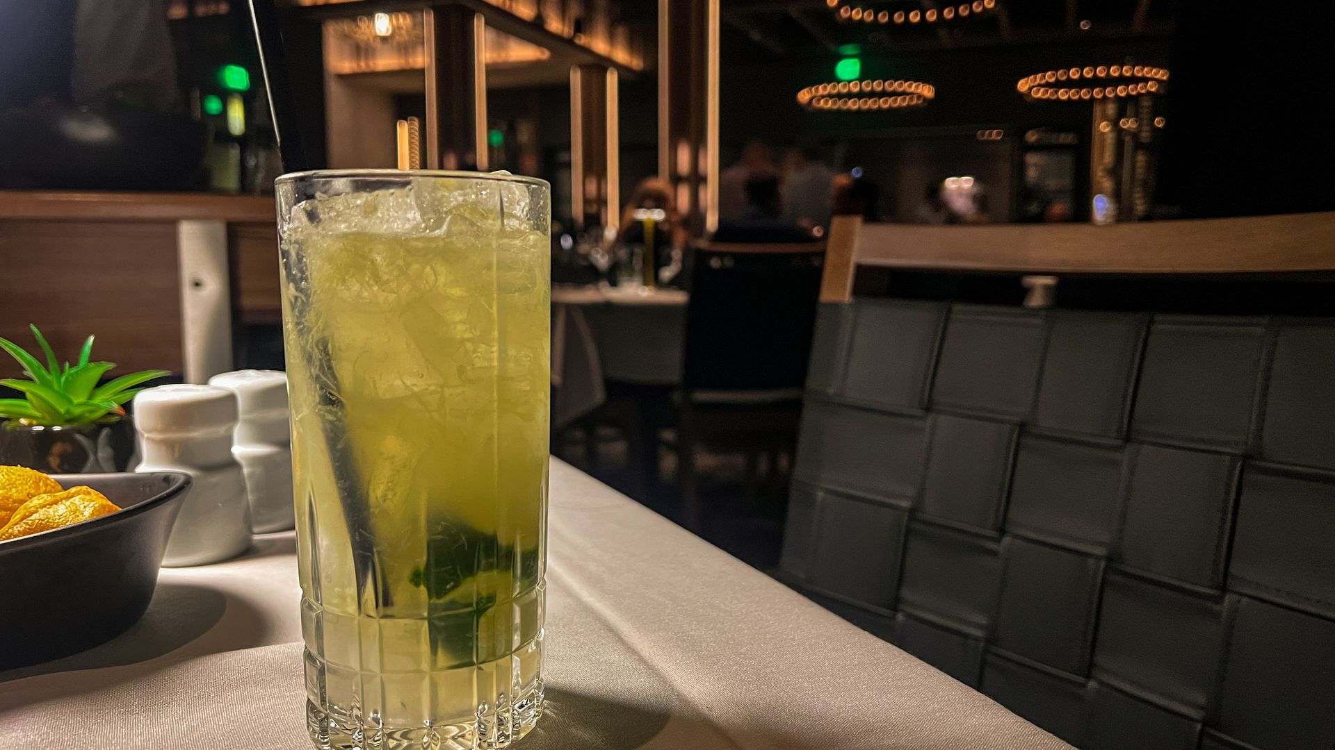 A tall glass filled with a light-colored drink sits on a table draped with a white linen.