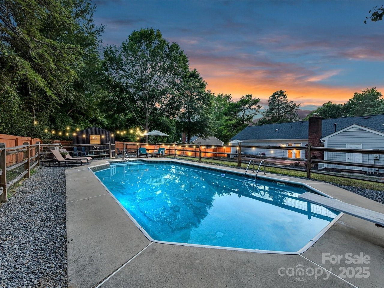 Outdoor swimming pool at dusk with blue water, concrete deck, lounge chairs, string lights, trees, and a house in the background under a colorful sunset sky.