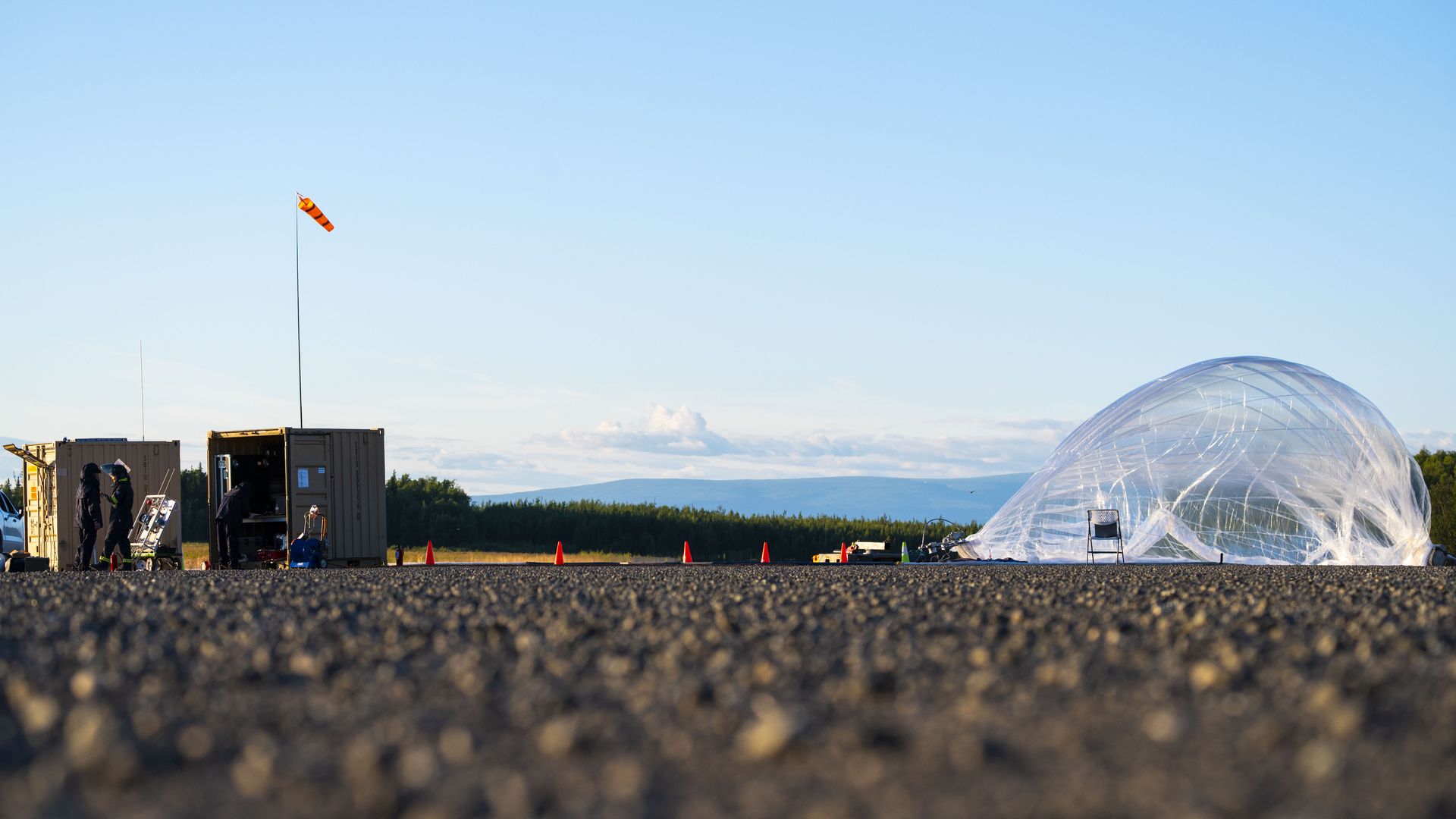 Low-angle view of an outdoor setup with two people near beige containers, an orange windsock, and a large inflatable, translucent plastic structure on gravel ground under a clear blue sky.