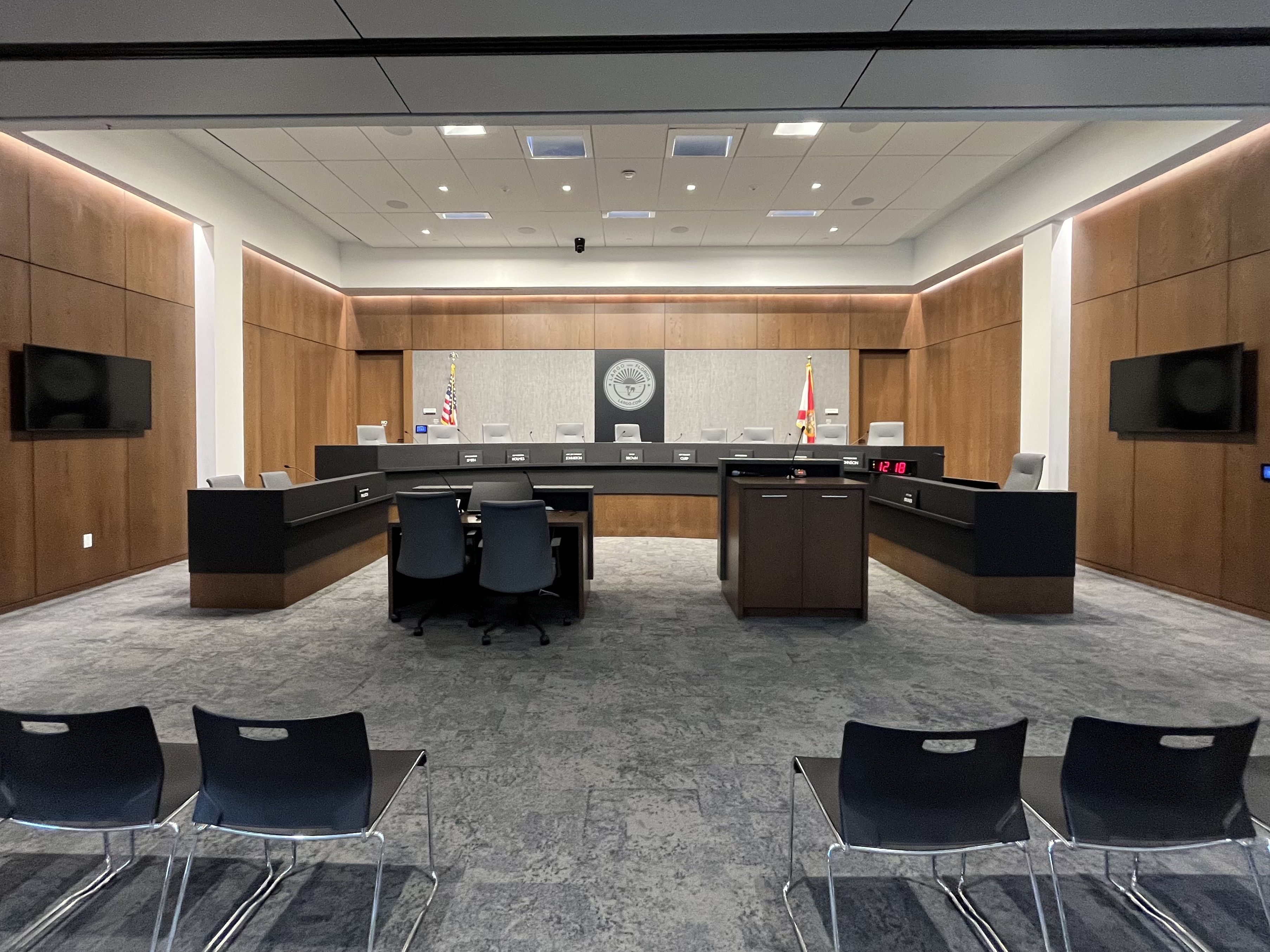 Interior of a modern courtroom or city council chamber with wood-paneled walls, a long central dais, gray backdrop with a circular seal, two flags (U.S. and Florida), and several chairs and monitors.