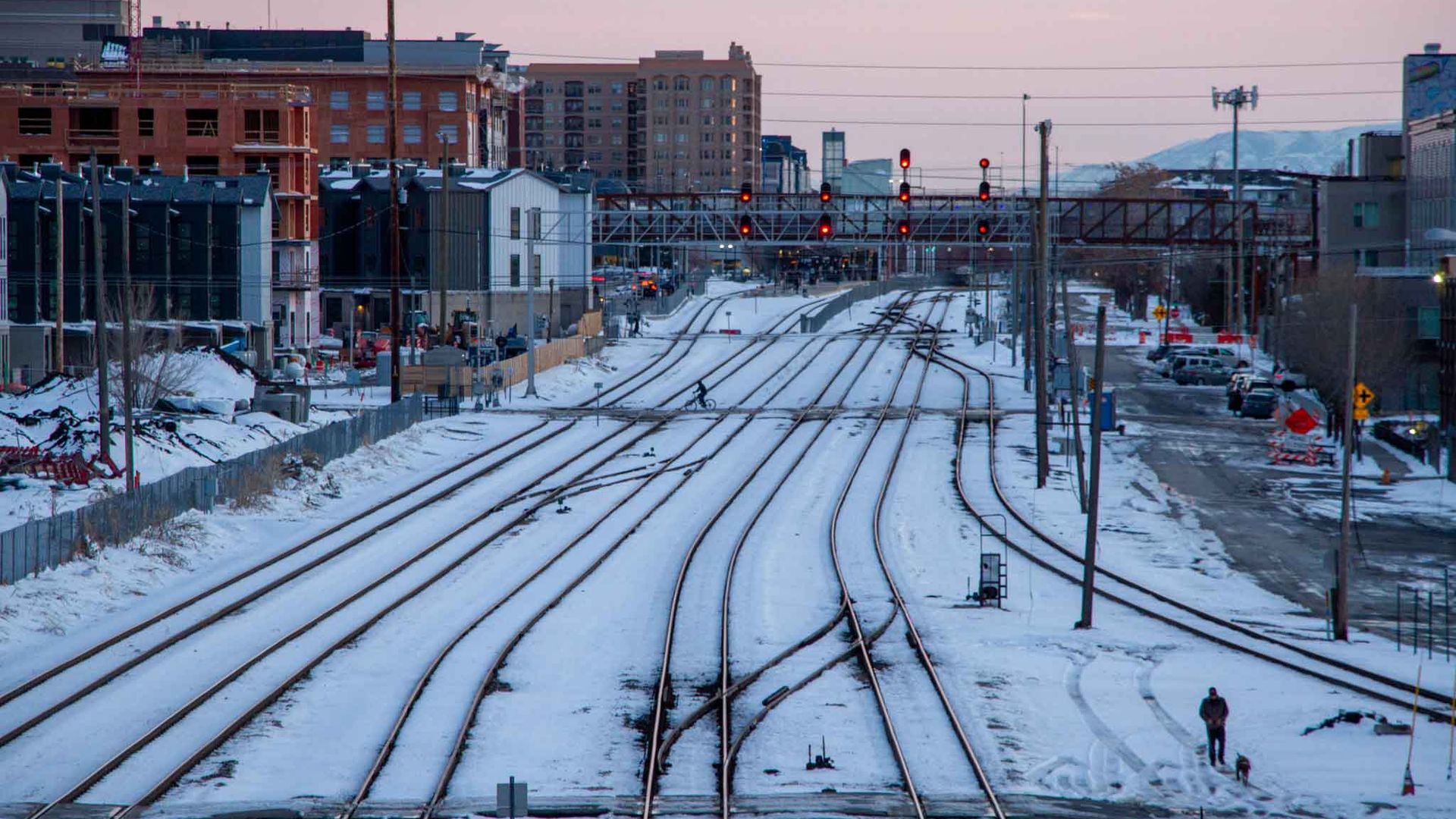 A man and dog walk between a snowy rail yard between two city neighborhoods.