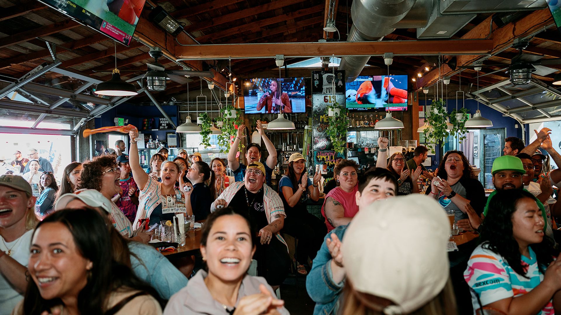 A crowded bar filled with diverse, cheering patrons at tables, some raising hands. TV screens hang overhead, showing a performance; exposed wooden ceiling, plants, and warm lighting.