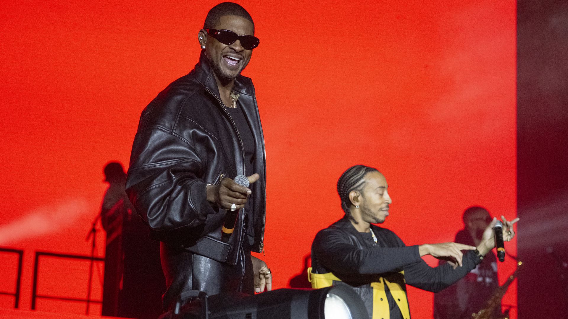 Two musicians perform in front of a solid red screen and gesture smiling toward the audience. A person plays a saxophone in the background. 
