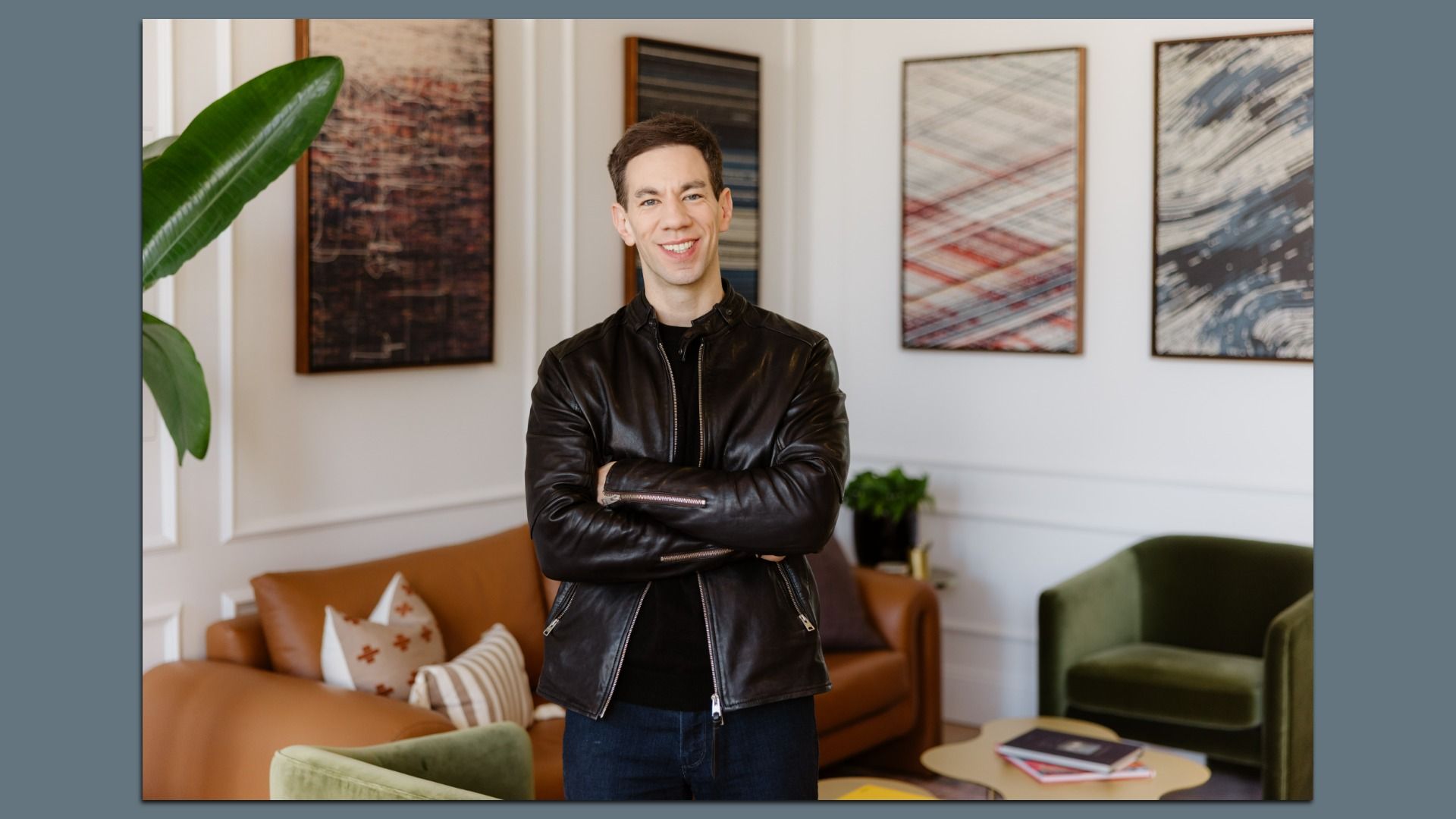 Smiling man in a black leather jacket stands with arms crossed in a modern living room, with abstract framed art on the wall, a large green plant, and a brown sofa in the background.