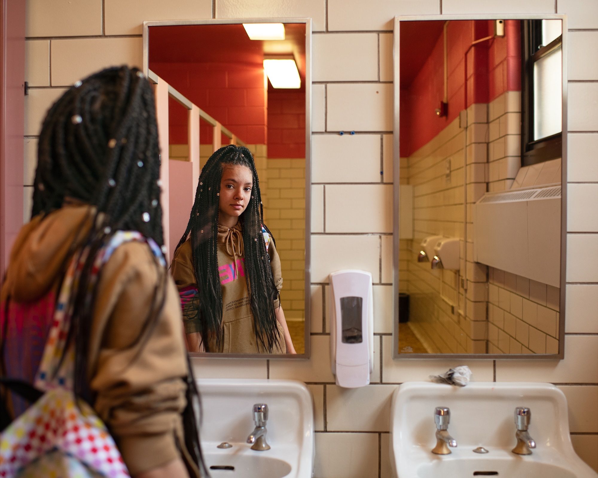Teen girl with long braided hair wearing a brown hoodie and colorful backpack looking at herself in a bathroom mirror with beige tiles and pink walls.
