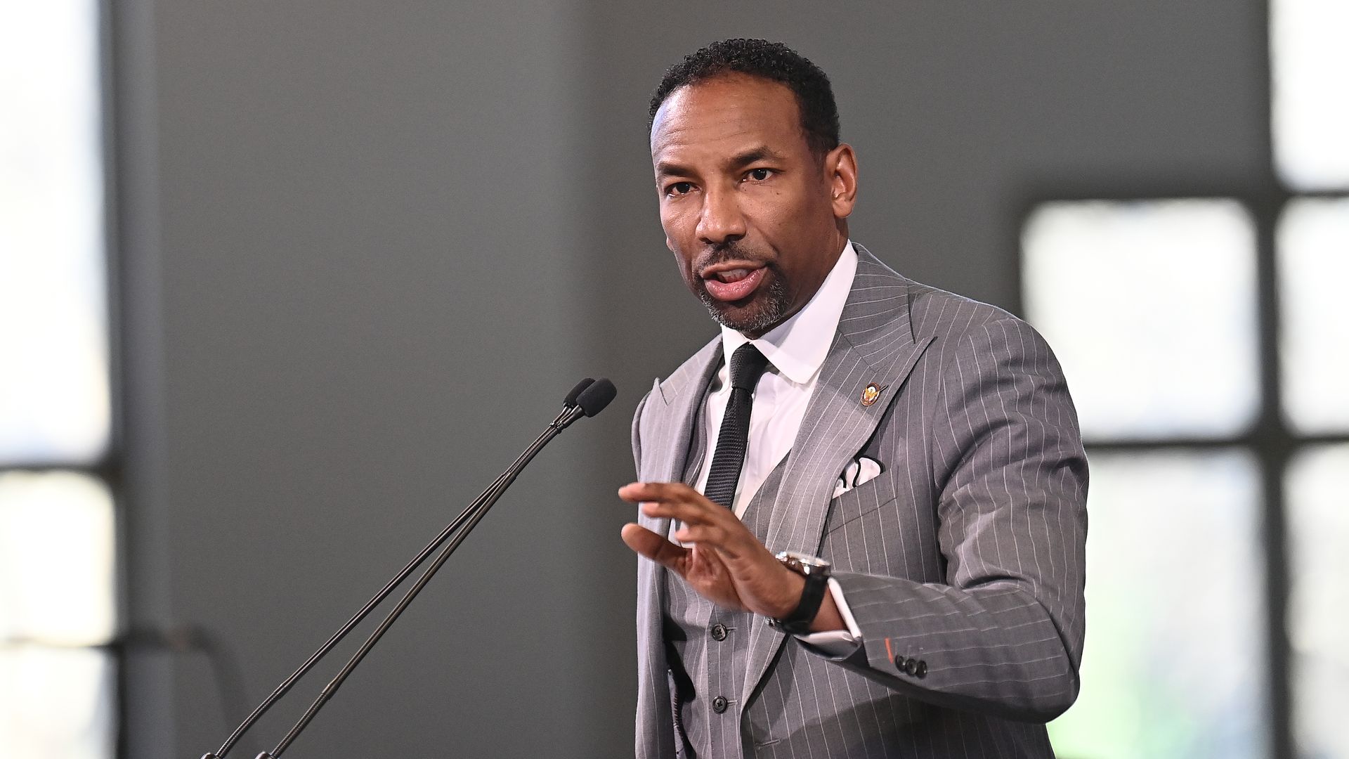 A man wearing a gray pinstripe suit, white collared shirt and dark-colored tie speaks at a microphone attached to a podium, with his left hand raised mid-air.