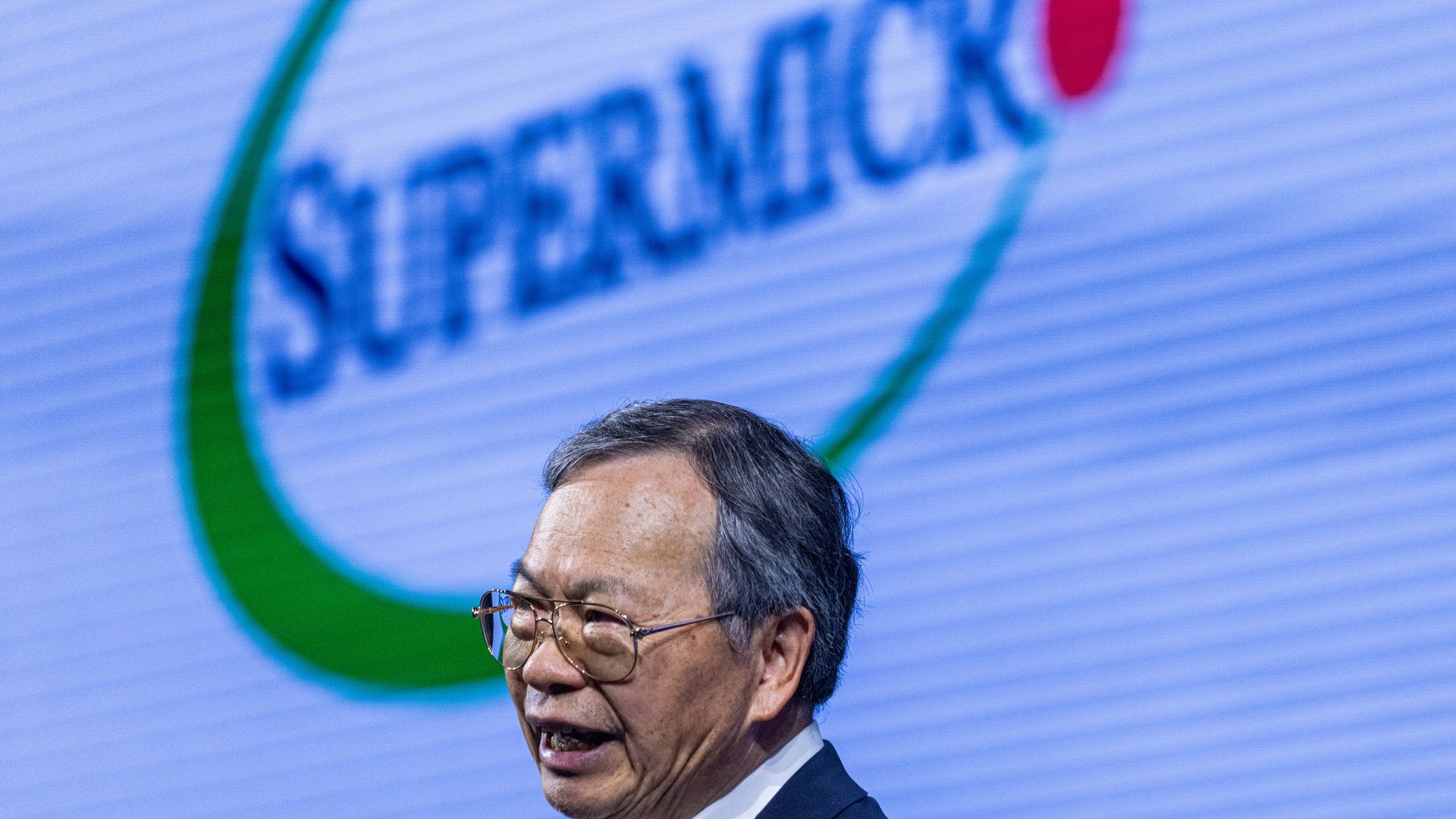 A man in a dark suit and glasses speaks into a lapel mic on stage, with a blue backdrop showing a large green circular logo behind him.