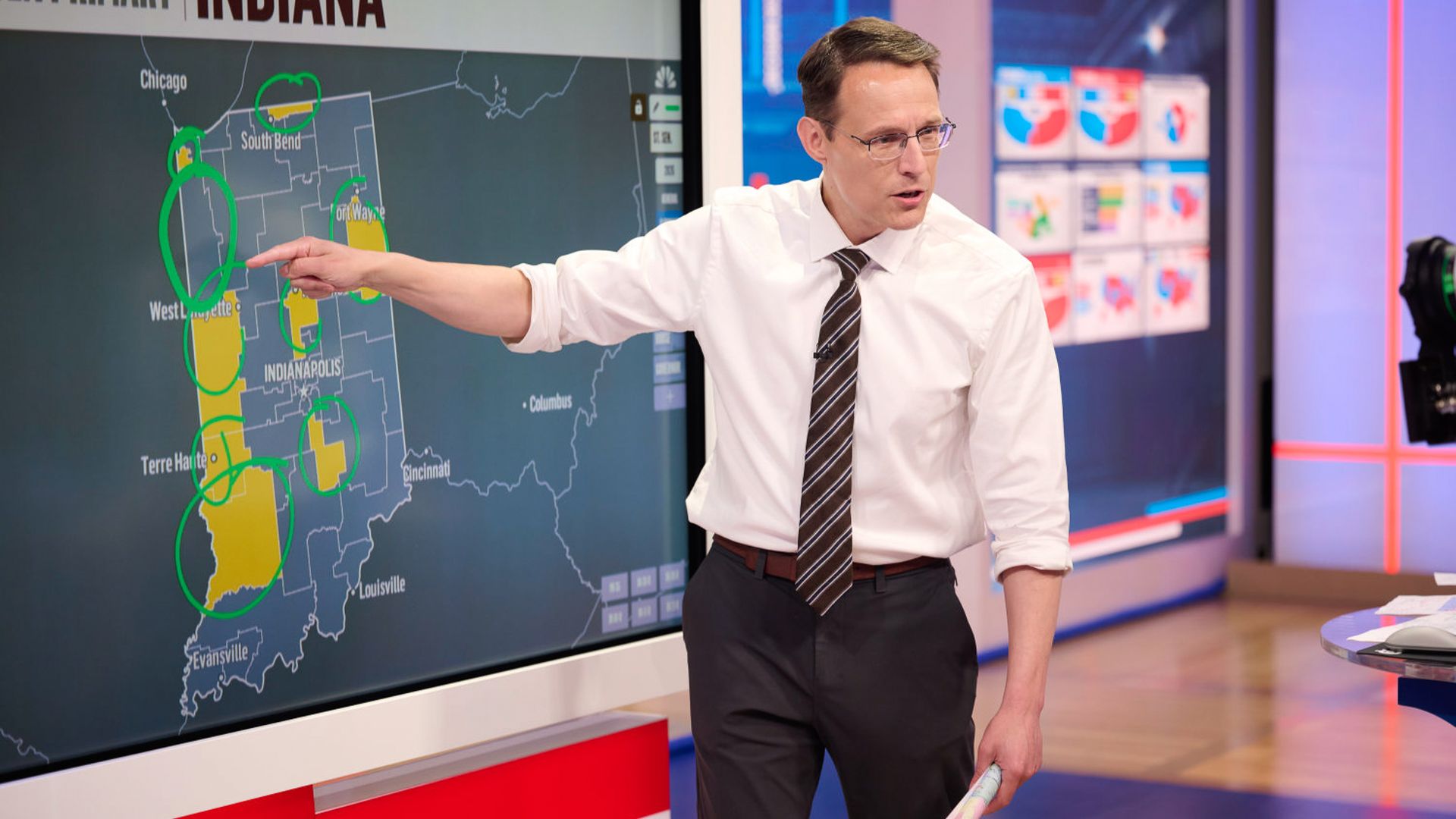 News anchor in a white shirt and striped tie points to a large touchscreen map of Indiana, with green circles around Chicago, South Bend, Indianapolis, Terre Haute, and West Lafayette; he holds a rolled paper.
