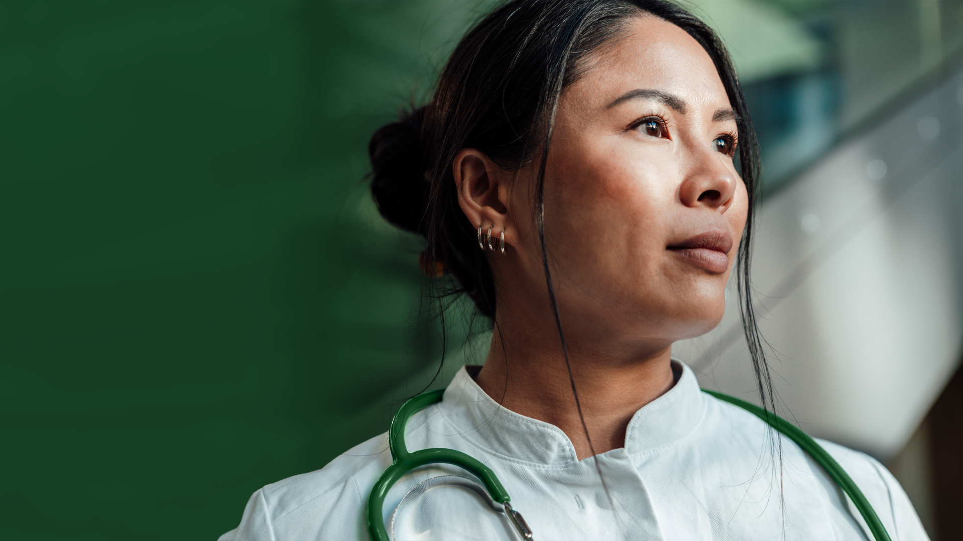 Close-up of a female healthcare worker with dark hair, wearing light scrubs and a green stethoscope, looking upward and to the right; blurred green background.