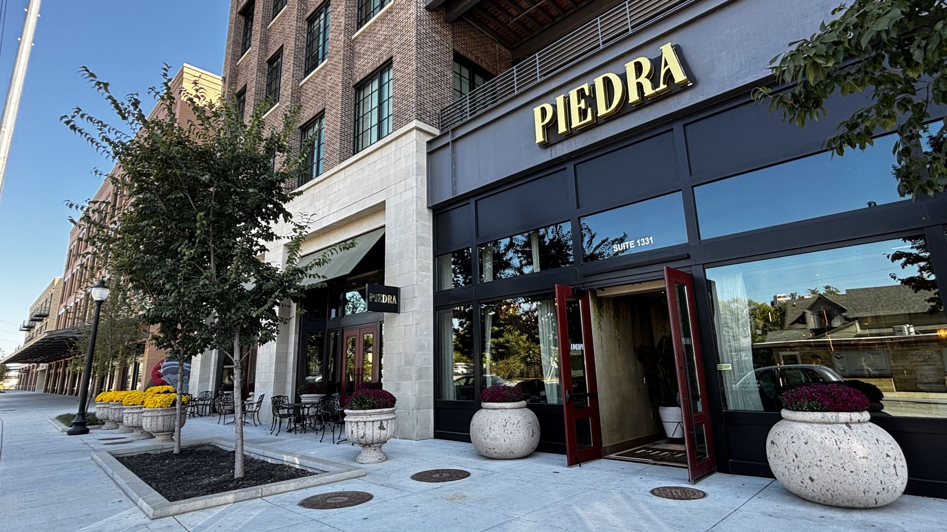 Sidewalk view of Piedra restaurant with open red doors, large beige planters with flowers, brick building, trees, chairs, and clear blue sky.