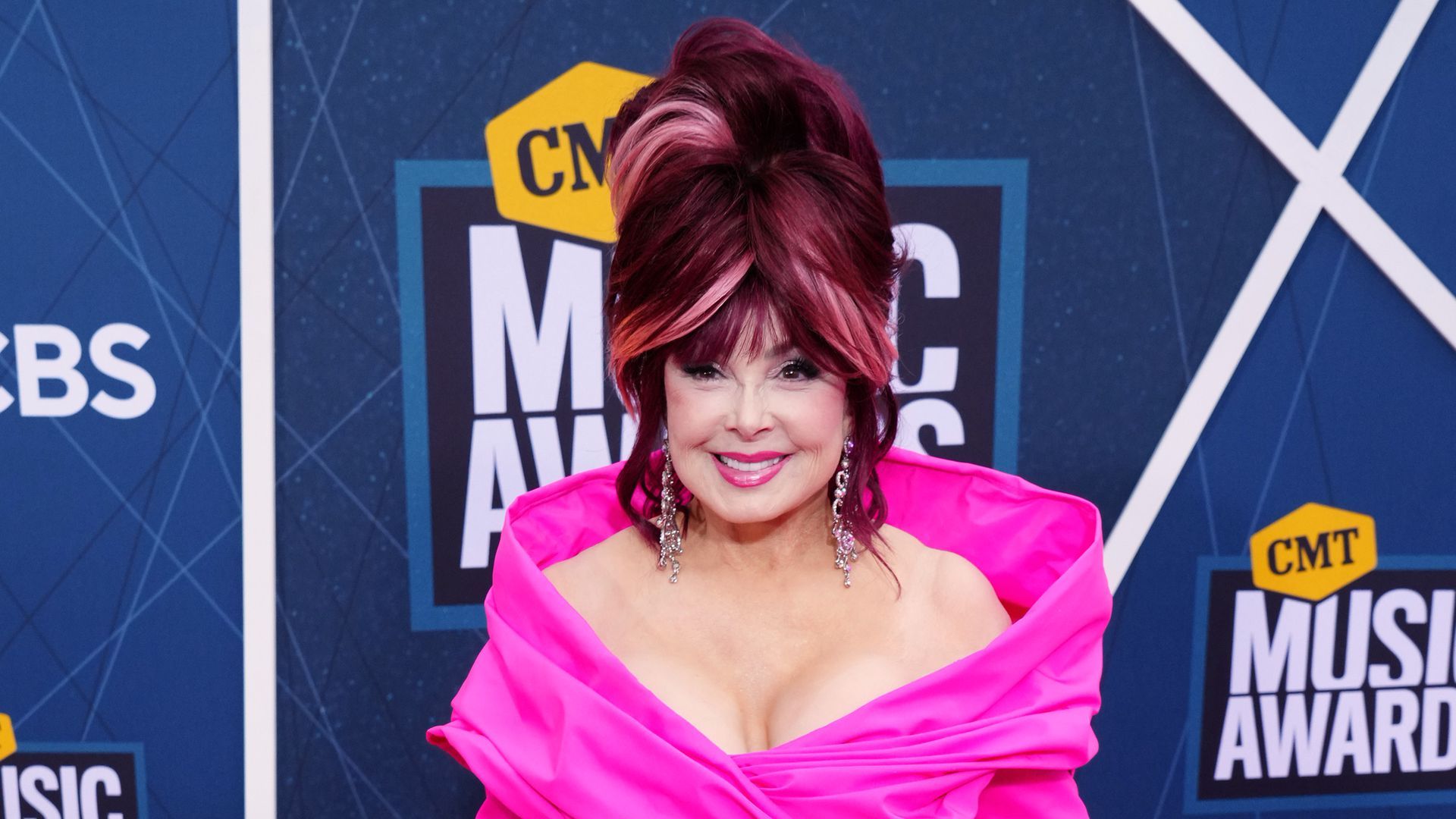 portrait of Naomi Judd with big hair and a pink dress in front of a sign that says "CMT Music awards"
