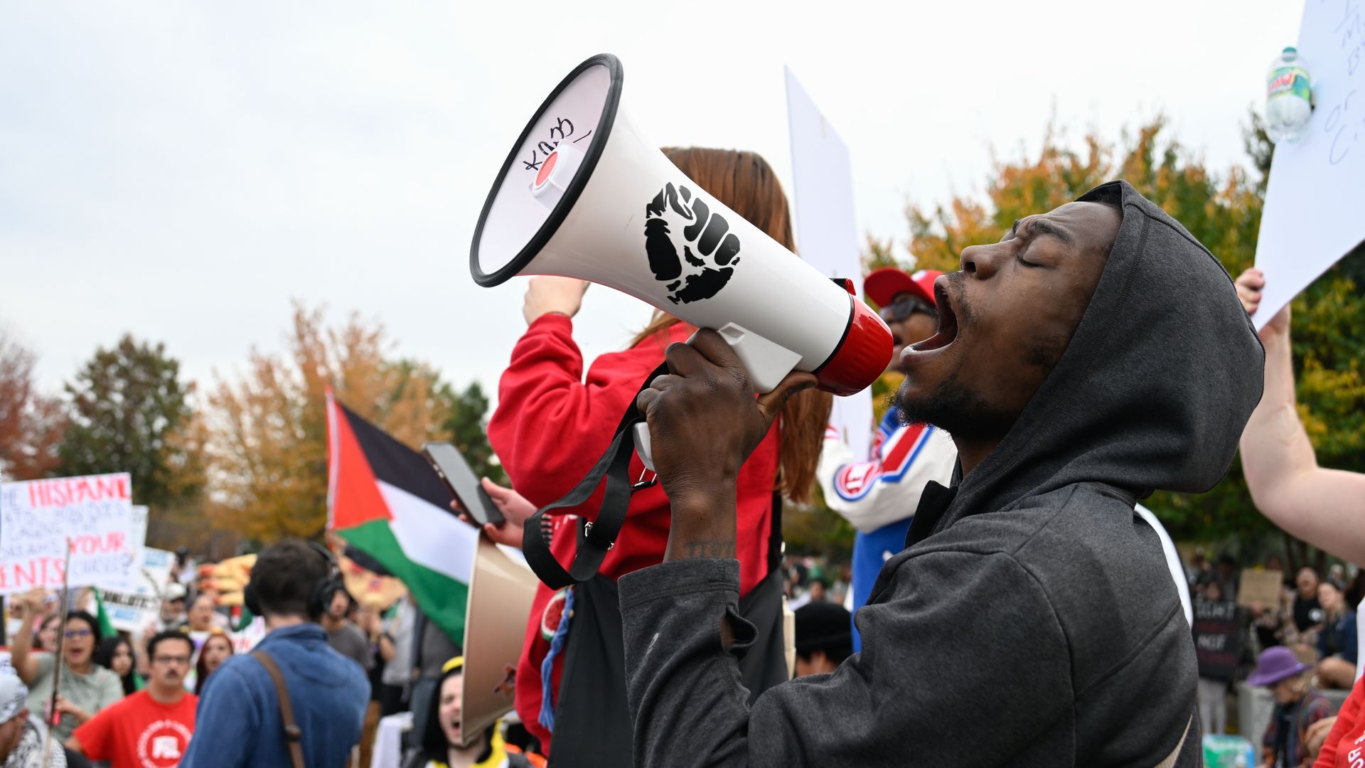 A person wearing a black hoodie shouts into a megaphone marked with an illustration of a closed fist. Dozens of protesters join a demonstration behind him.