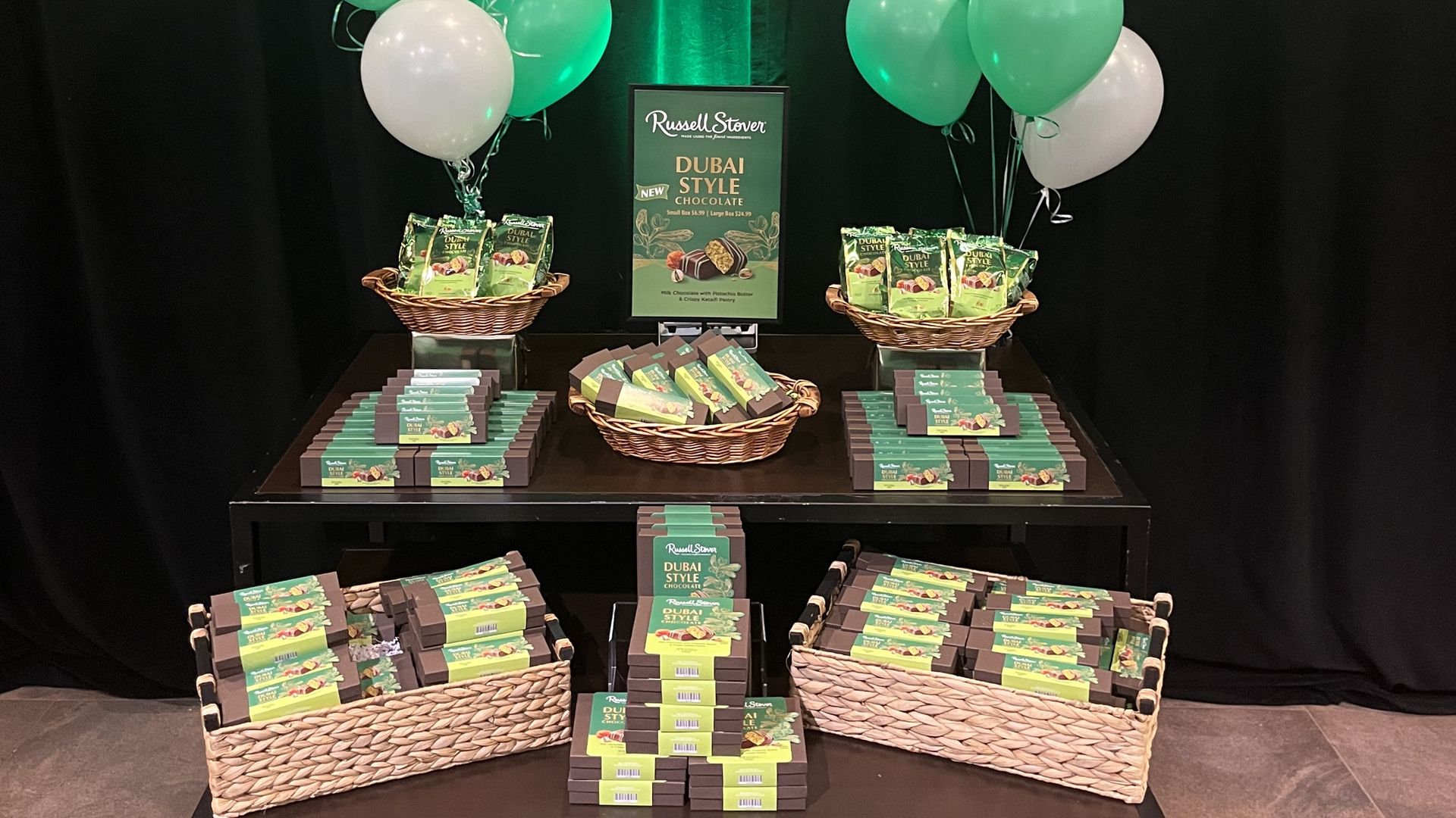 Display of Russell Stover Dubai Style chocolate products in green and brown packaging on black table with green and white balloons and promotional sign.