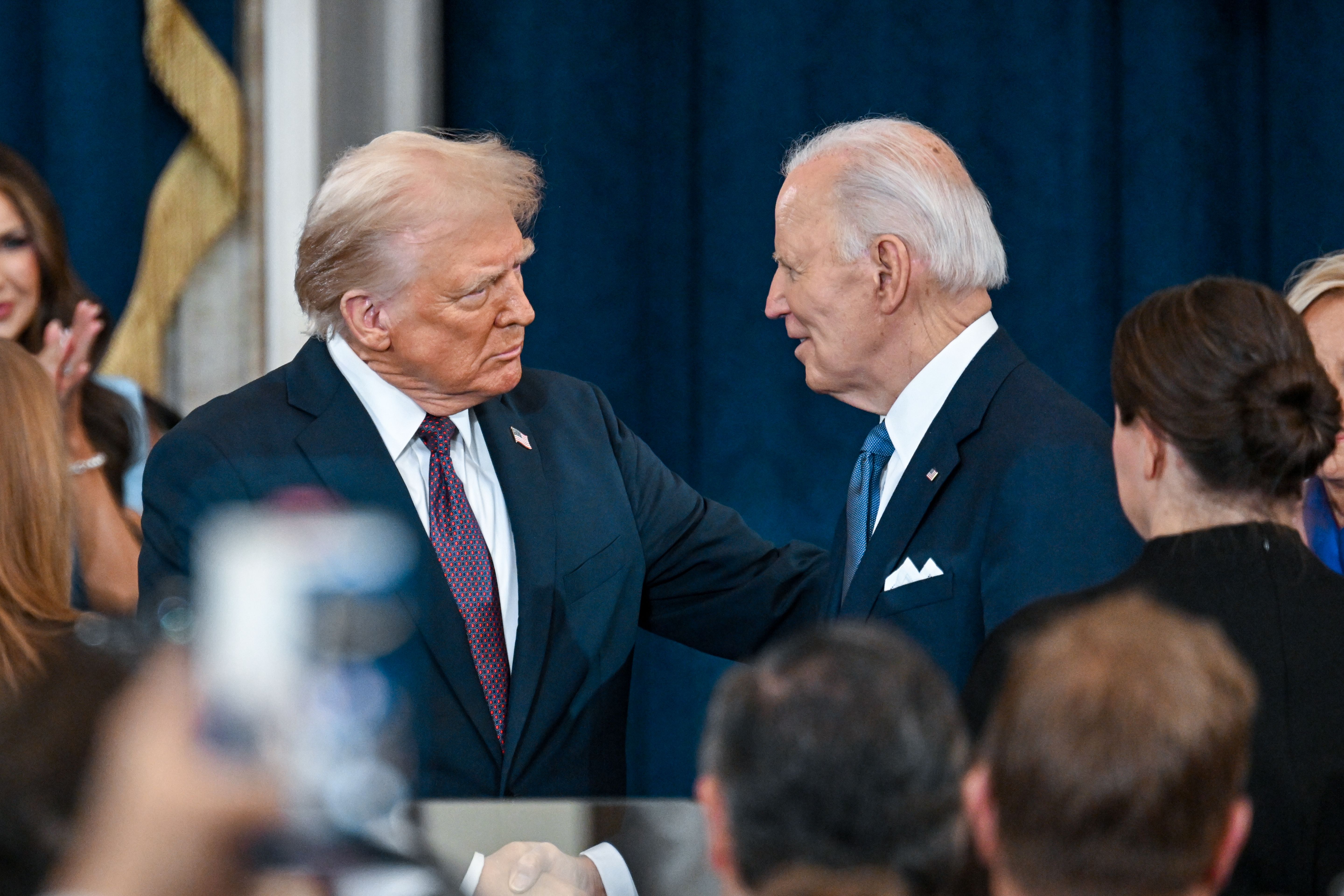 Two elderly men in formal suits stand facing each other in a crowded room. One is gesturing with his hand on the other's shoulder, while the other listens attentively. The background includes people clapping, and the setting appears formal, with a dark curtain visible.
