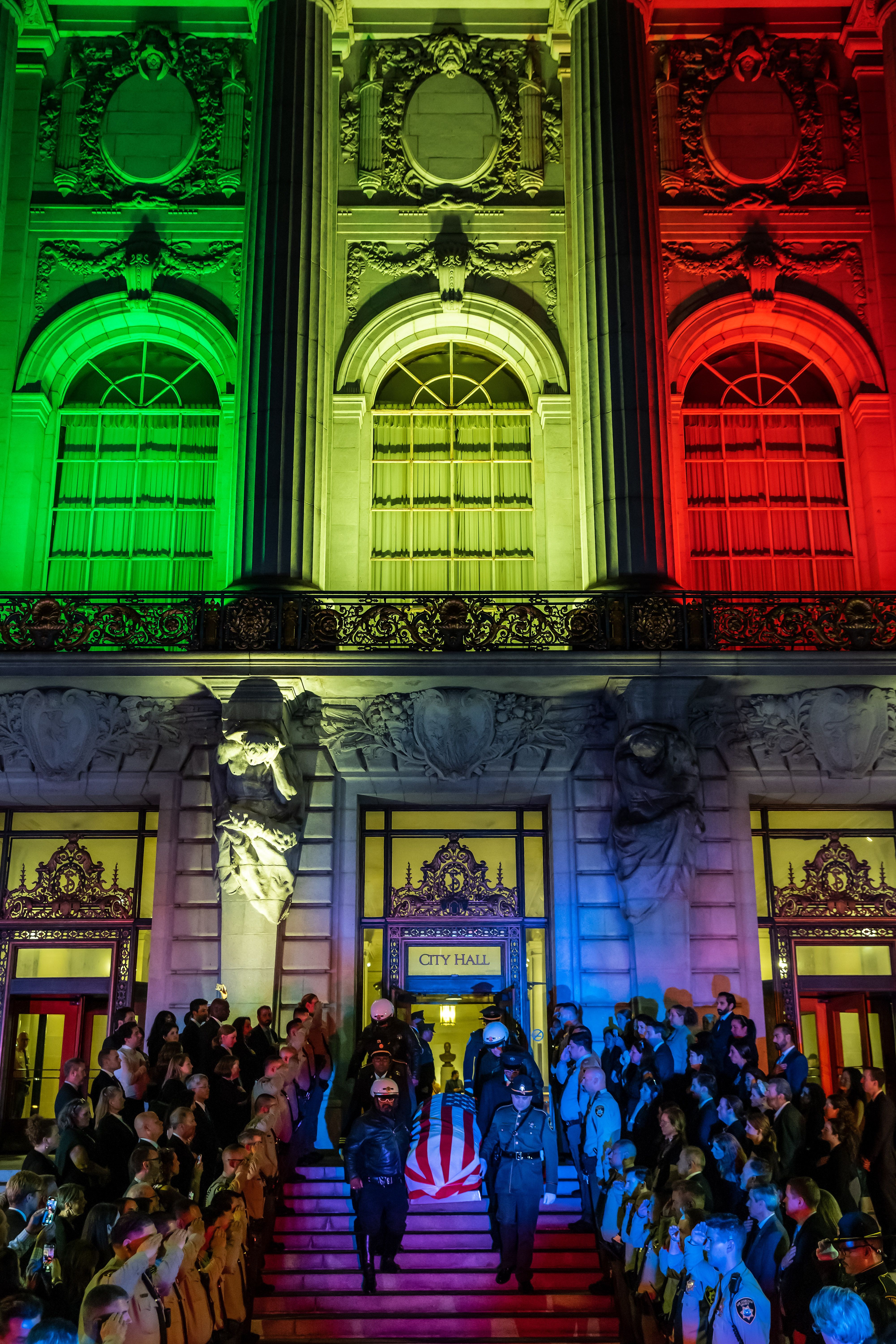 Lights on San Francisco City Hall shine green, yellow and red as honor guard members carry down Feinstein's casket with a crowd overlooking.