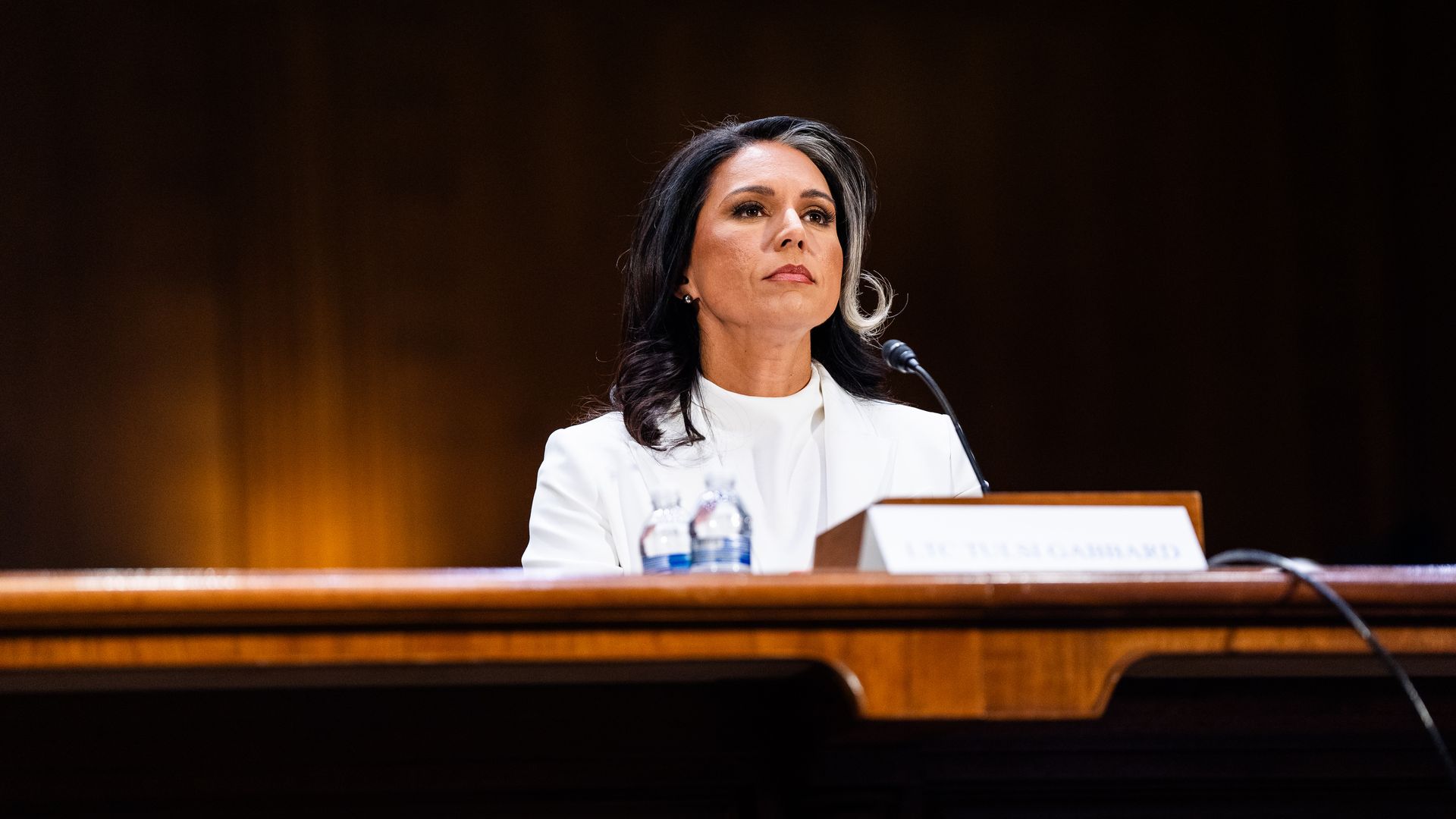 Tulsi Gabbard, in a white suit, sitting at a table with a microphone and large binder in front of her.