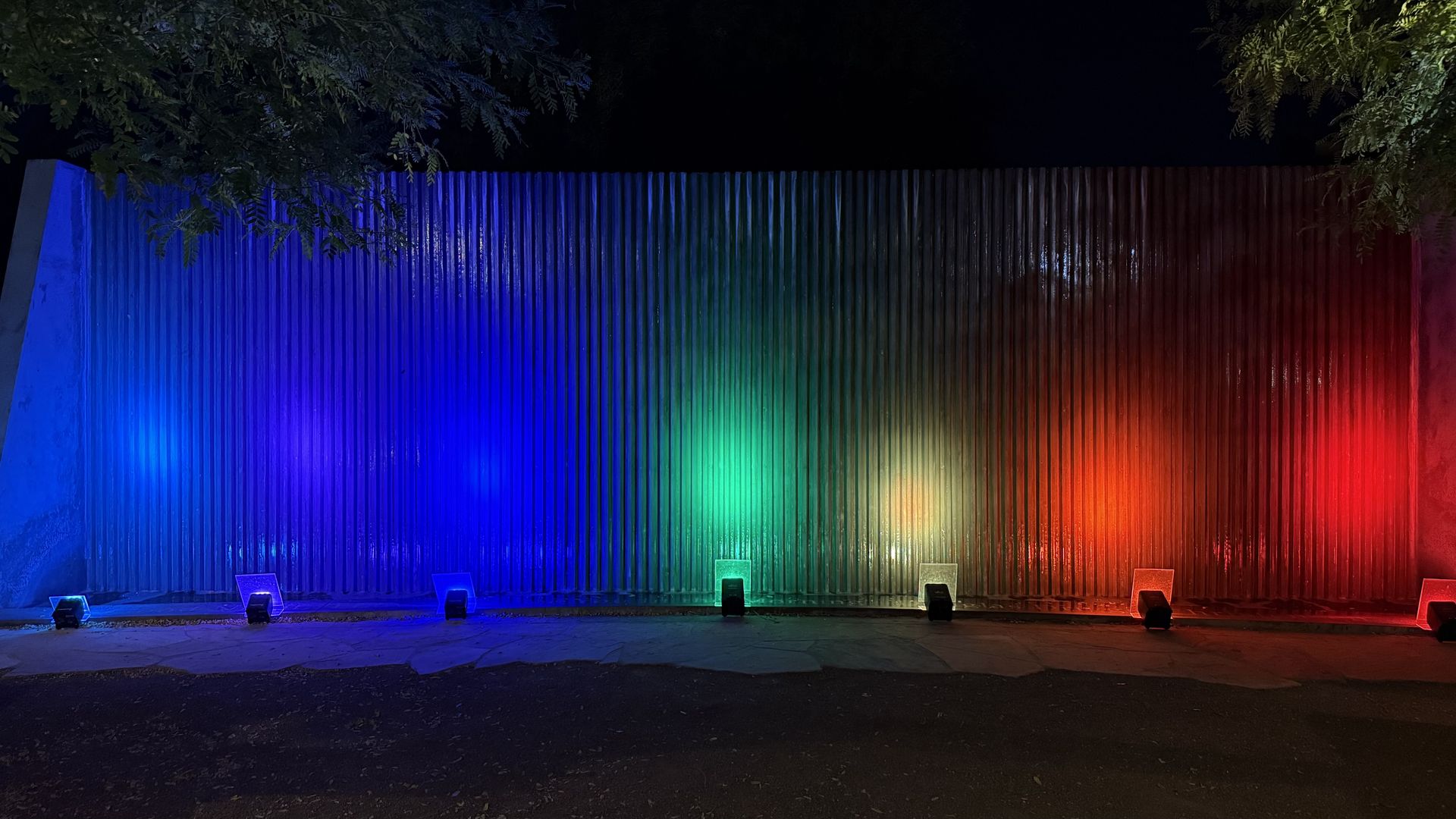 Night view of a corrugated metal wall lit by colorful ground lights in blue, green, yellow, orange, and red, with tree branches framing the top edges.