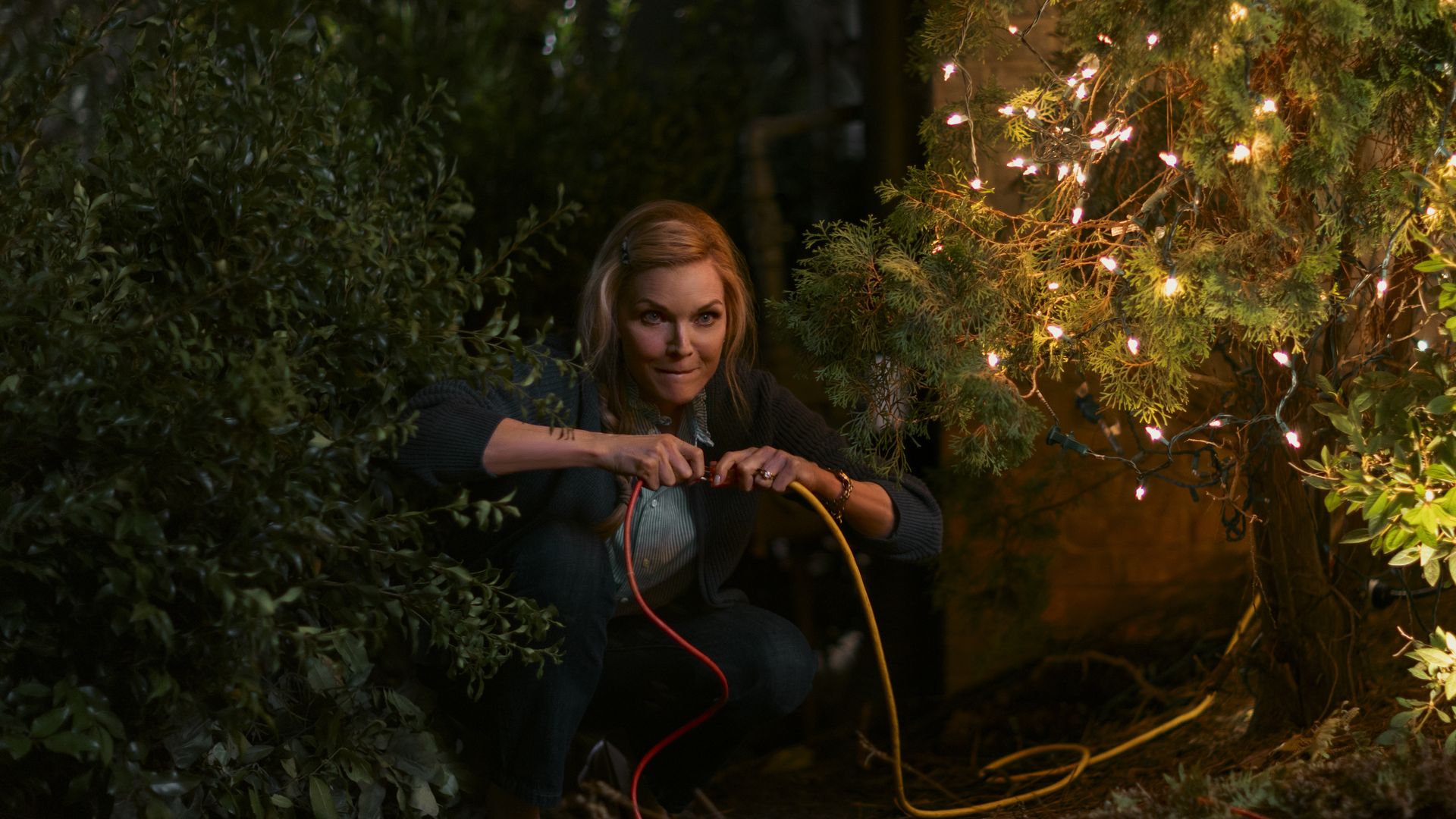 Woman crouches at night connecting red and yellow extension cords near a bush decorated with glowing string lights, with a focused expression on her face.
