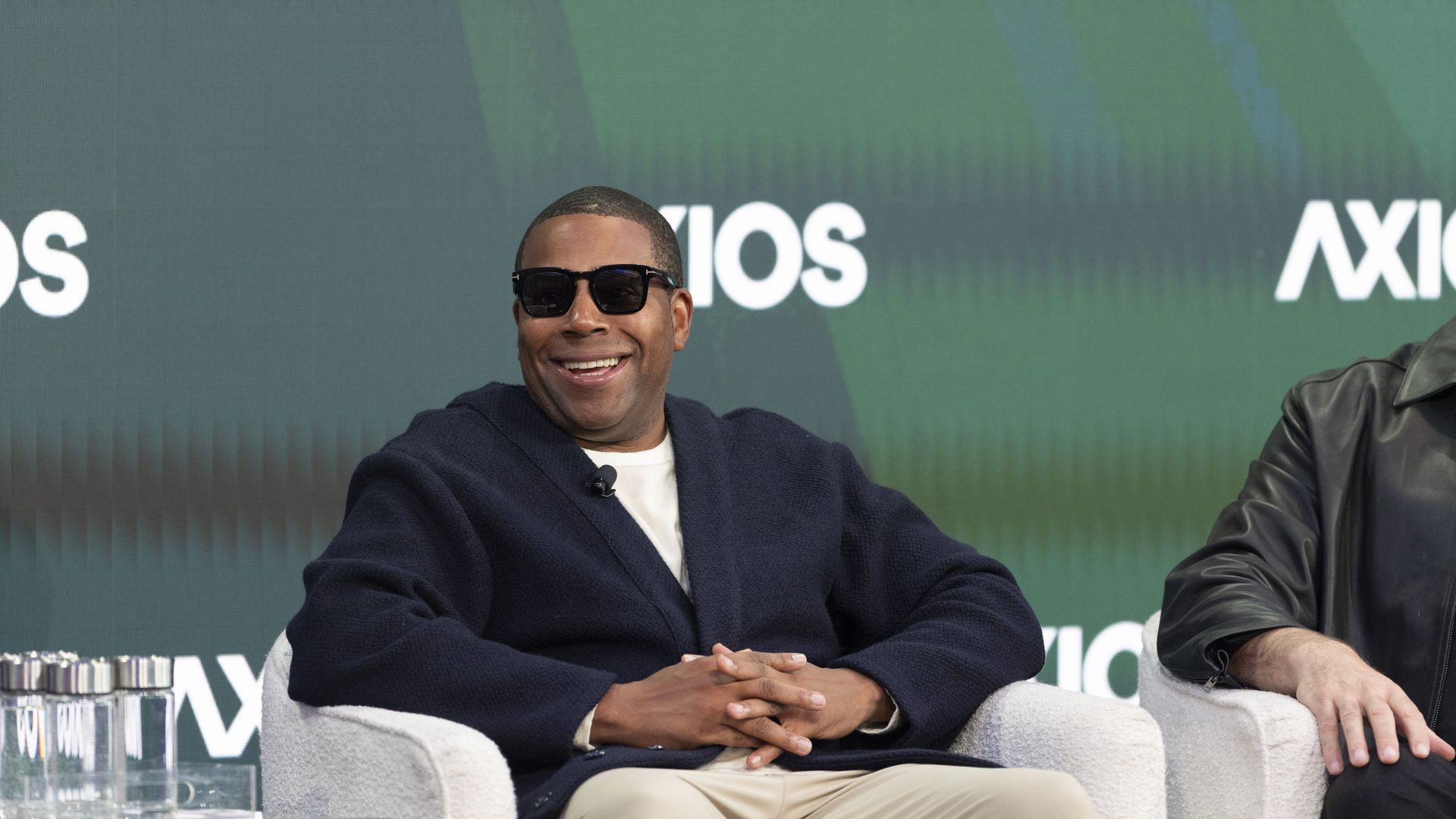 Smiling man in a navy blazer and sunglasses sits on a panel against a green Axios backdrop; hands folded in his lap, with a glass-topped table in front.