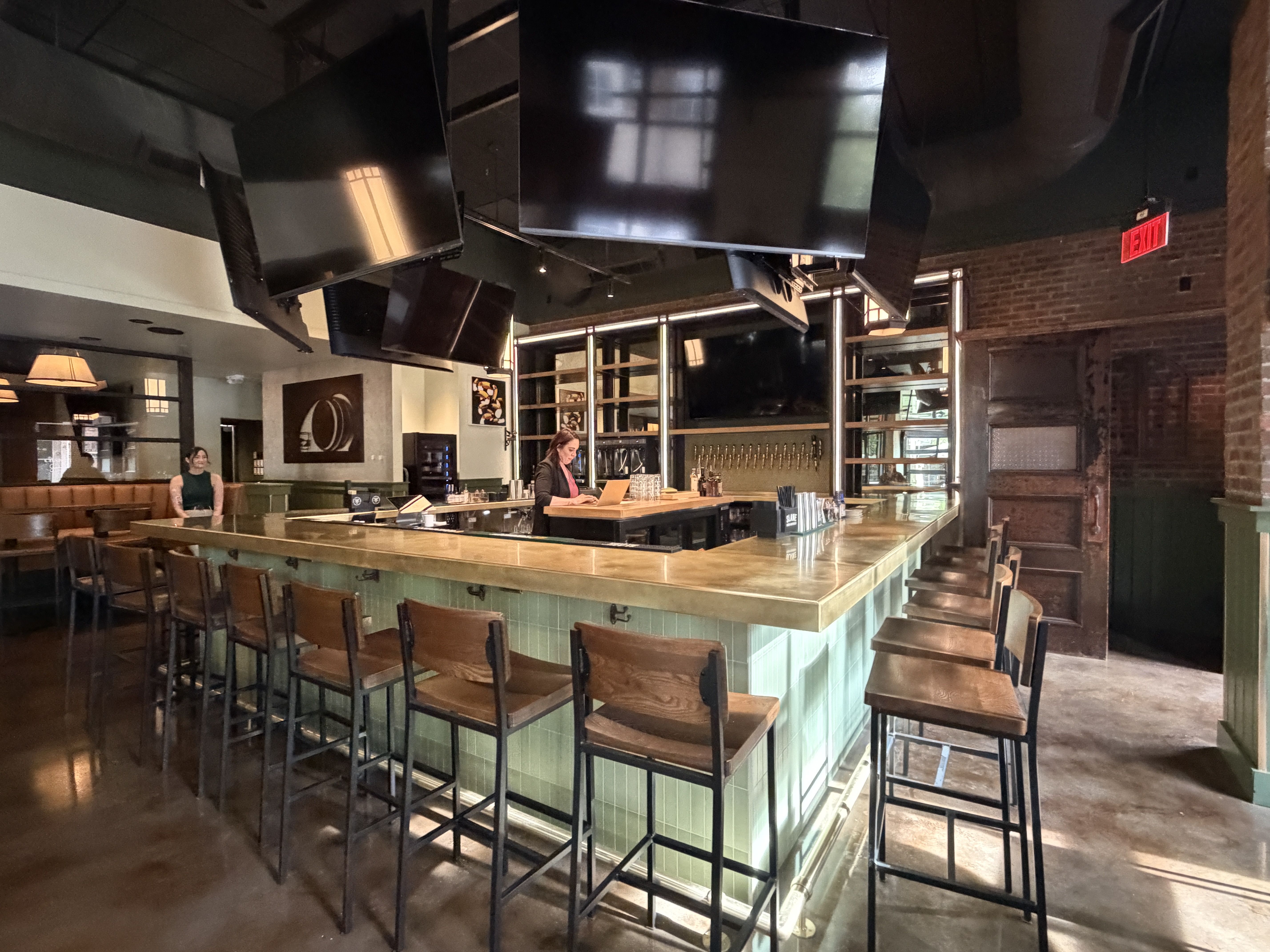 Bar interior with a mint-green tiled counter and wooden stools. A bartender uses a laptop behind the bar; a customer sits at the far end. Ceiling screens, brick walls, and an exit sign are visible.