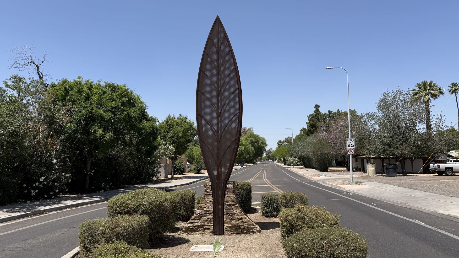 Tall, metal leaf-shaped sculpture with visible veins in the center median of a street, surrounded by small green shrubs.