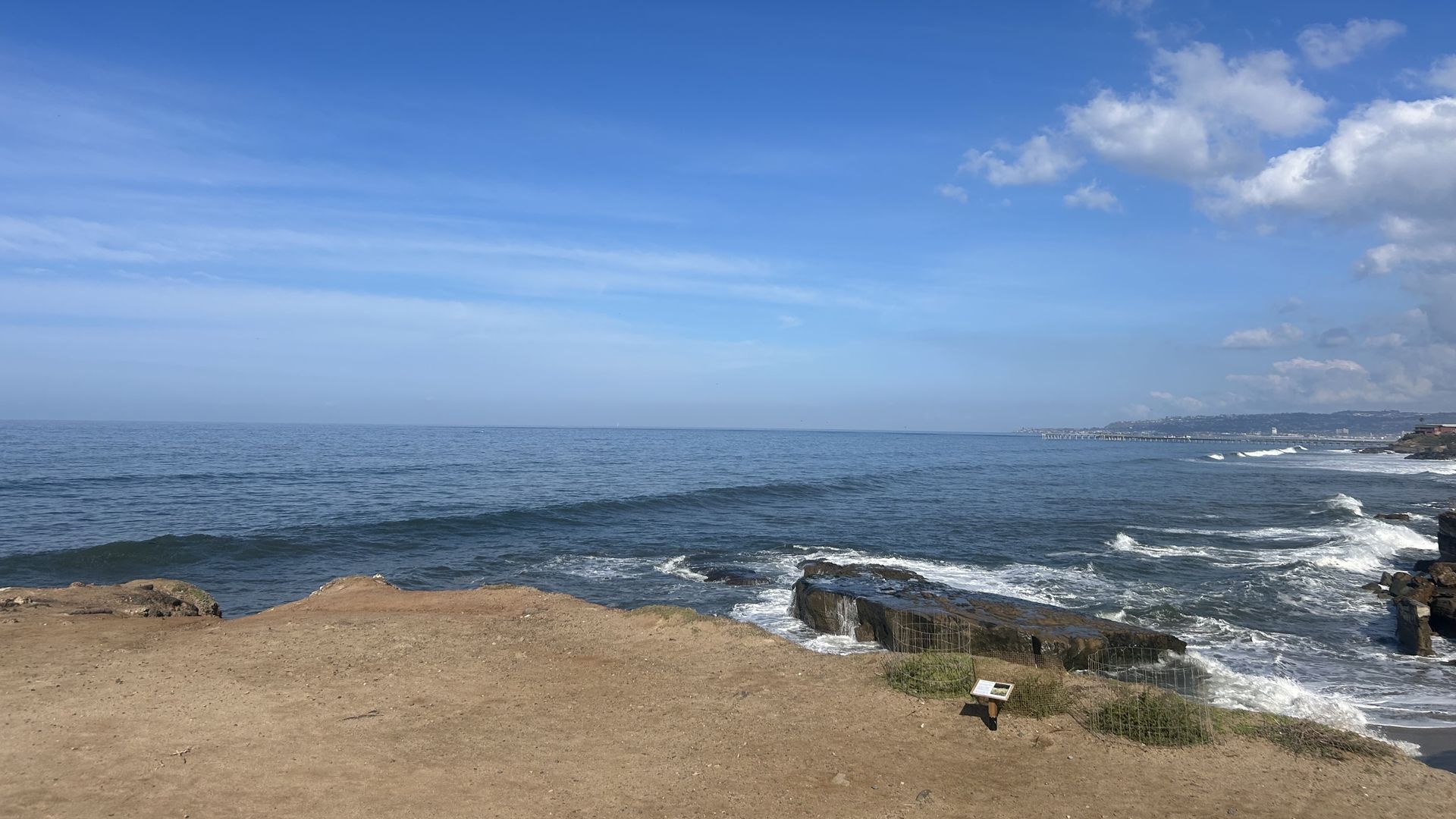 Wide ocean view under a deep blue sky; gentle waves lap a rocky, sandy shore. Right side shows clouds and distant coastline; a small sign sits on the sand near rocks.