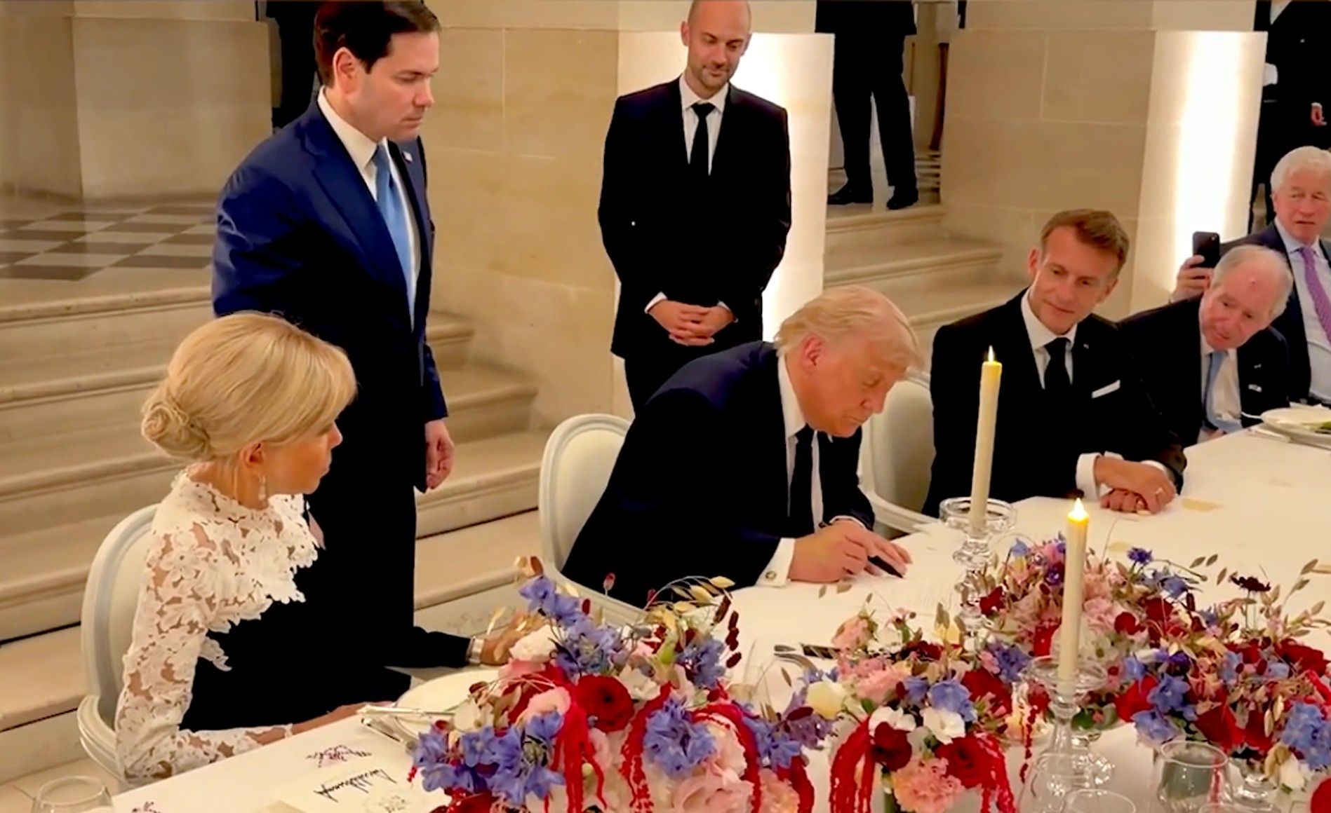 Formal dinner: a man in a dark suit signs a document at a long table with candles and a floral centerpiece; a blonde woman in white lace sits left; others in suits stand nearby in a hall.