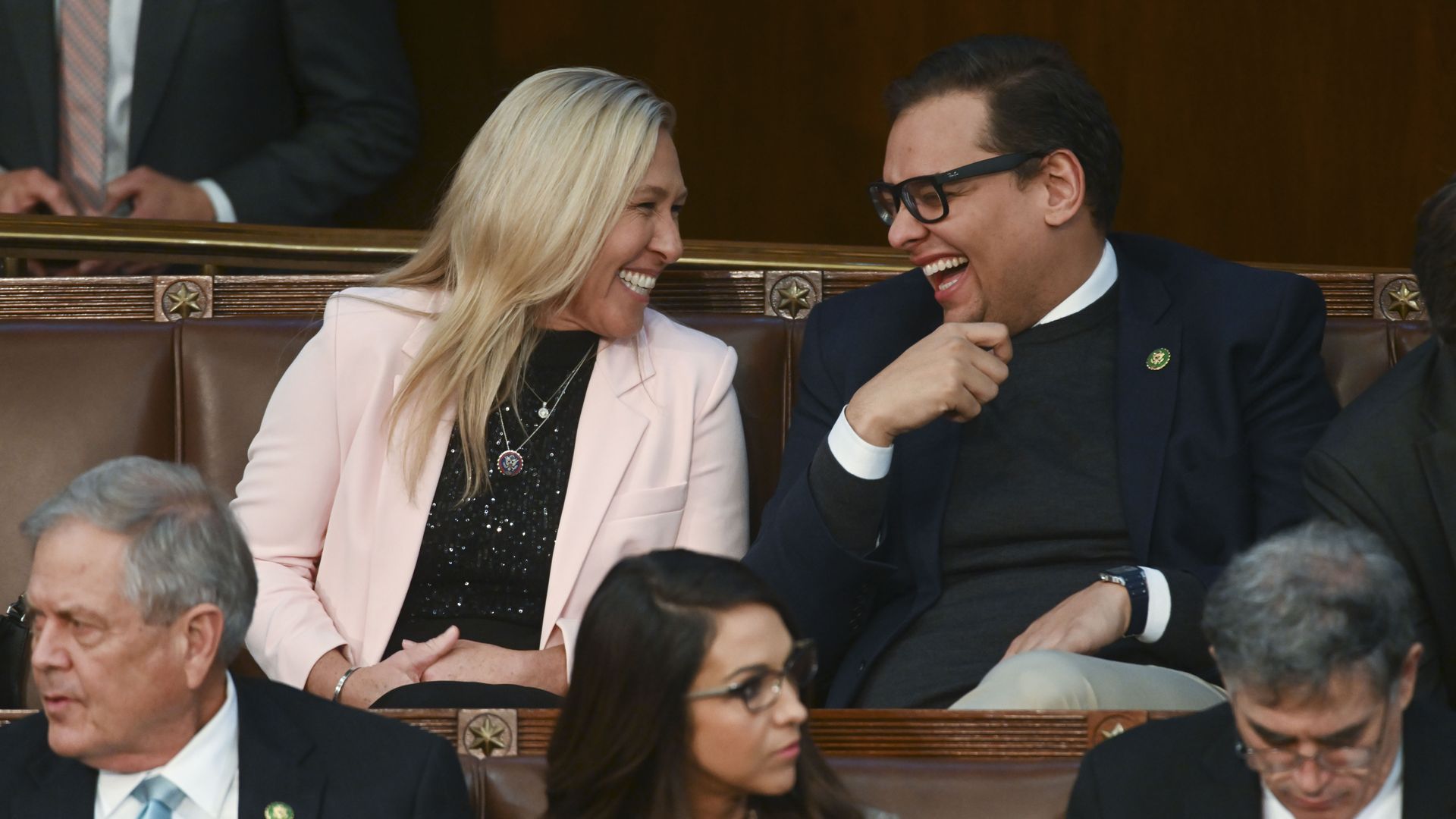 Two people in the top row laughing and talking, woman in a pink blazer and man in dark suit with glasses, seated on brown leather chairs with others around them in a formal setting.