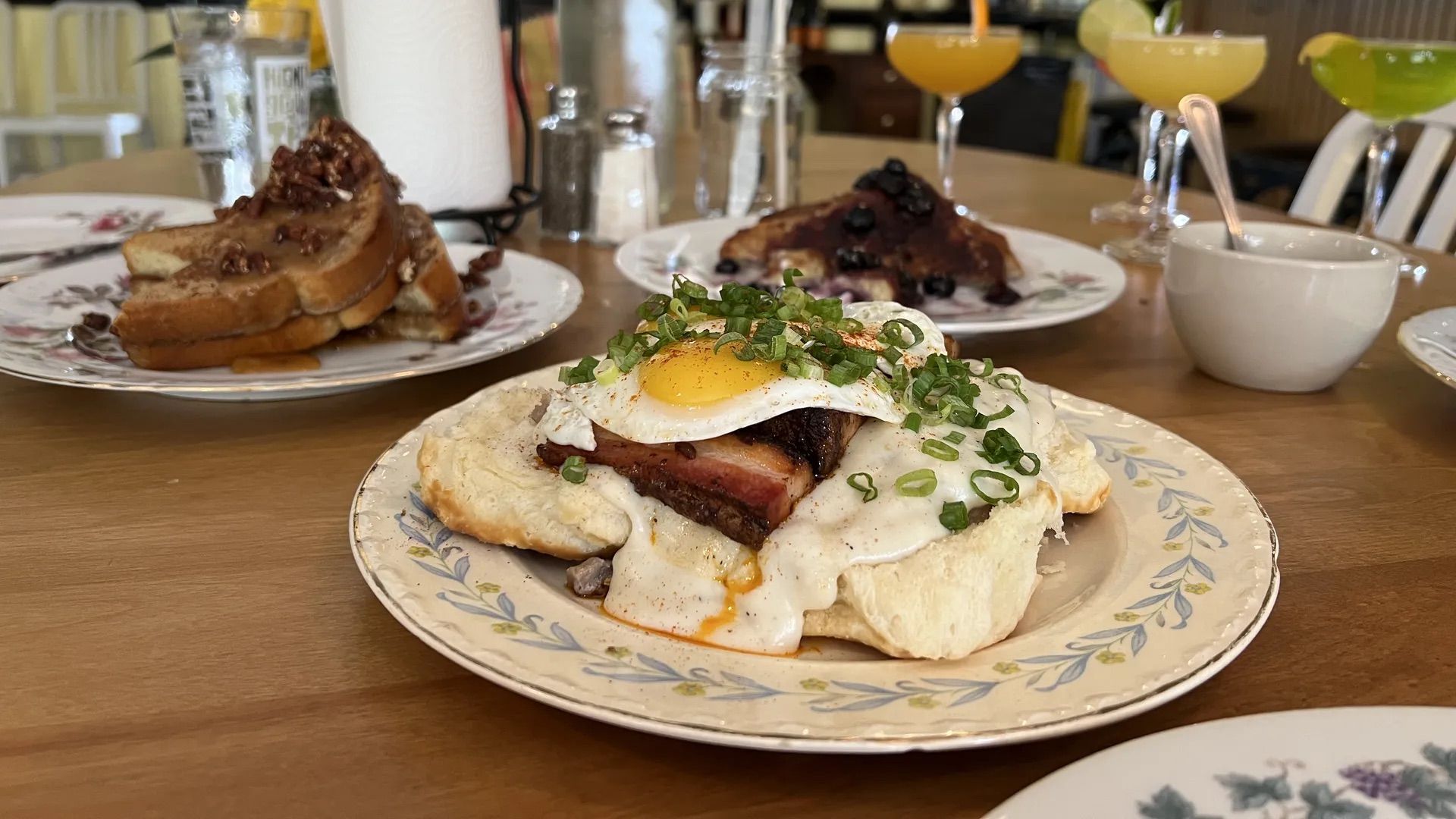 Plate with biscuit topped with thick ham, white gravy, a sunny-side-up egg, and chopped green onions on a wooden table with other plates and orange, yellow, and green drinks in the background.