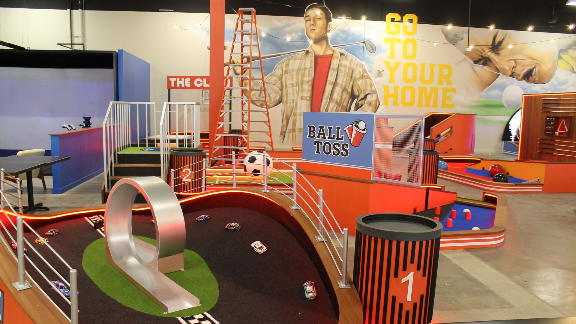 Indoor game center with an orange-themed miniature car racing track and ball toss game, featuring murals of a man with a golf club and a focused face with the text \