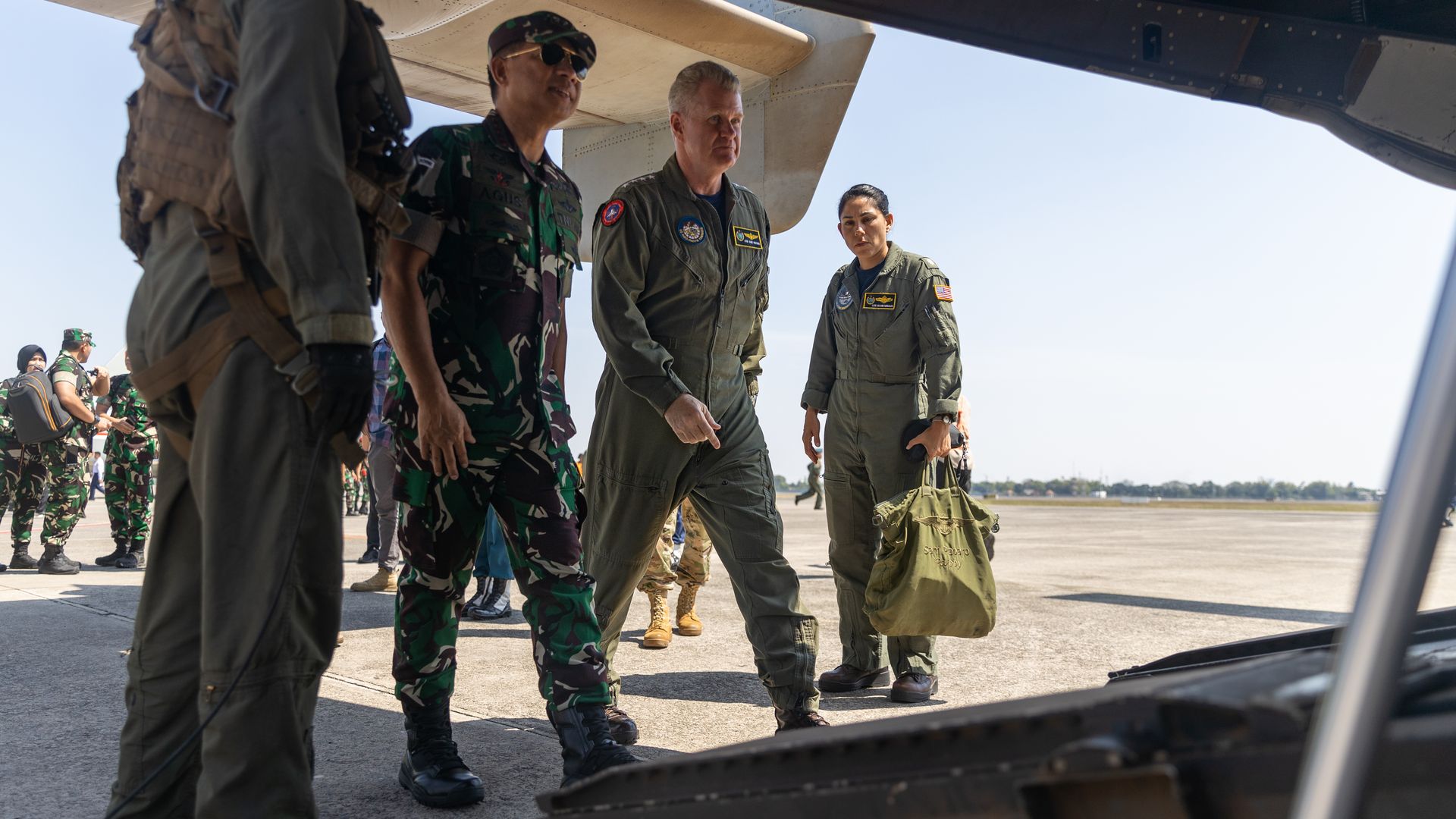 Three men and one woman in military uniform are seen. Two are walking onto an open cargo ramp of a plane. Blue skies are seen in the background.
