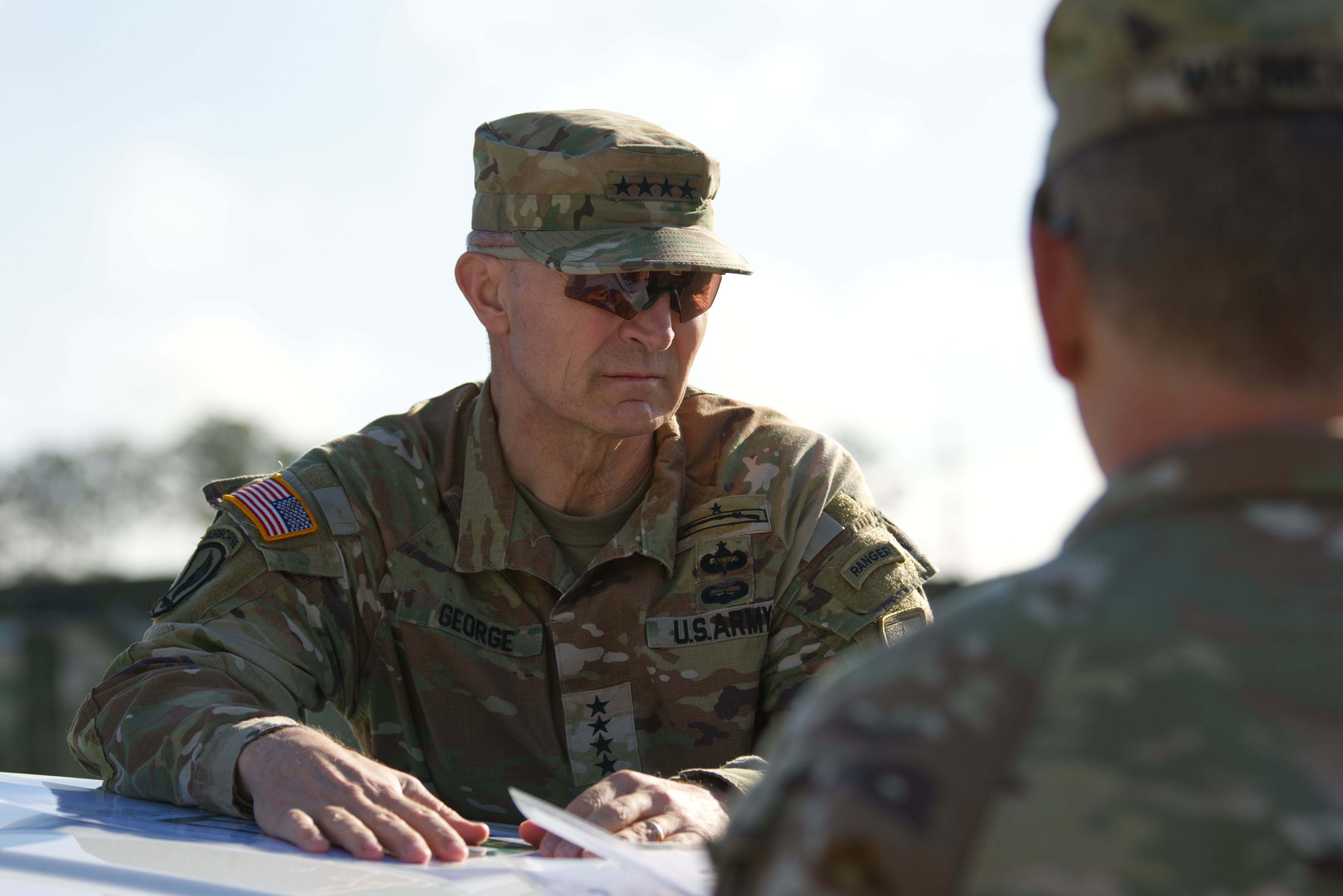 Two U.S. Army officials in camouflage talk outdoors. The man facing the camera wears a camo cap and sunglasses. Another man, in the foreground, has his back to the camera.