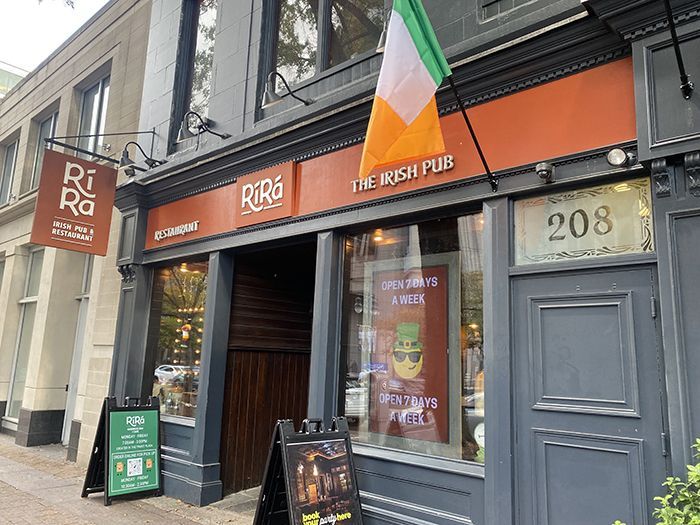 Gray-brick storefront for "Ri Ra" Irish Pub with an Irish flag above an orange sign reading "THE IRISH PUB." Window shows a poster and the text "OPEN 7 DAYS A WEEK"; address 208.