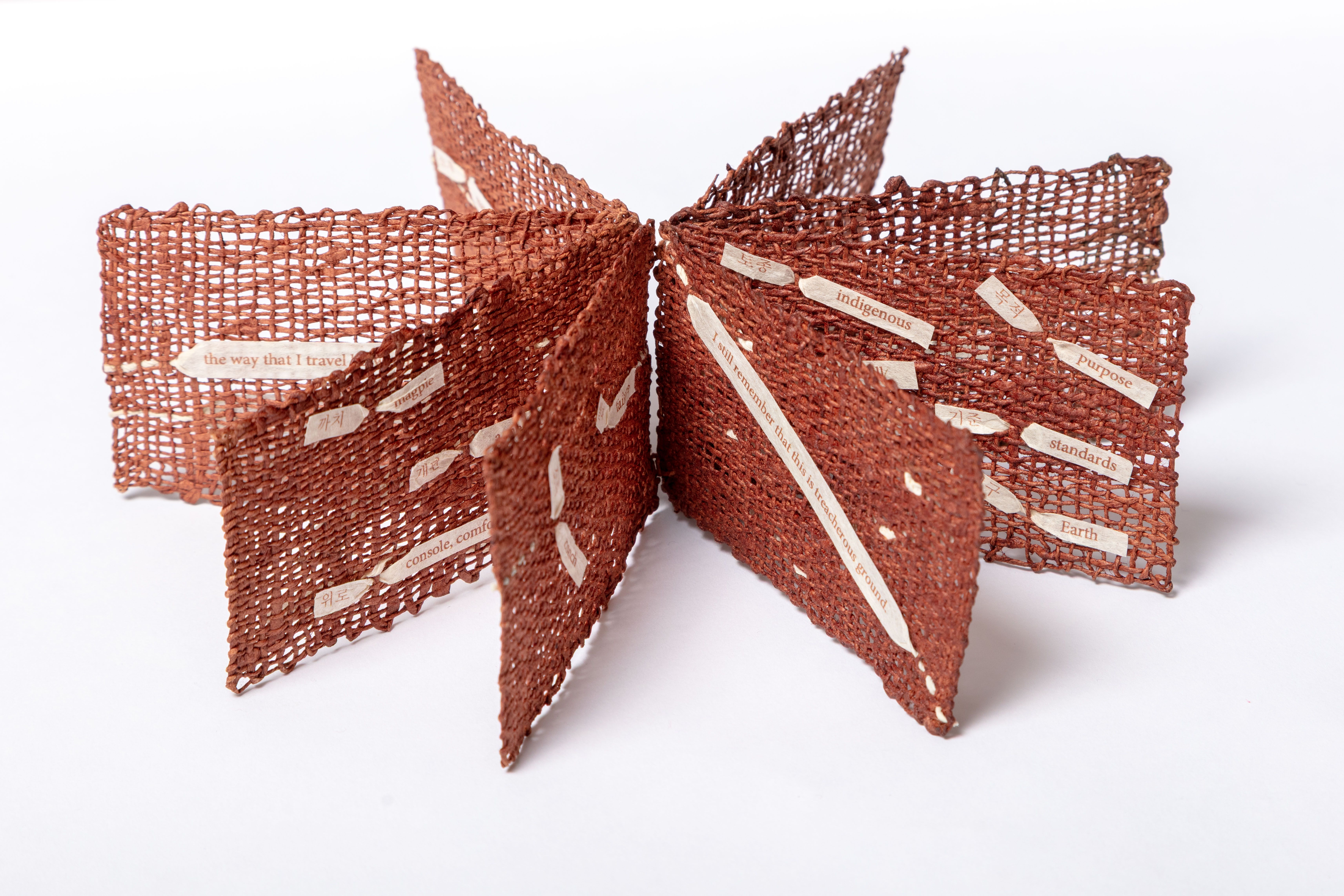 A large brown woven mesh bow against a white background, with small paper tags bearing words like "indigenous", "Earth", and "the way that I travel" attached to the ribbons.