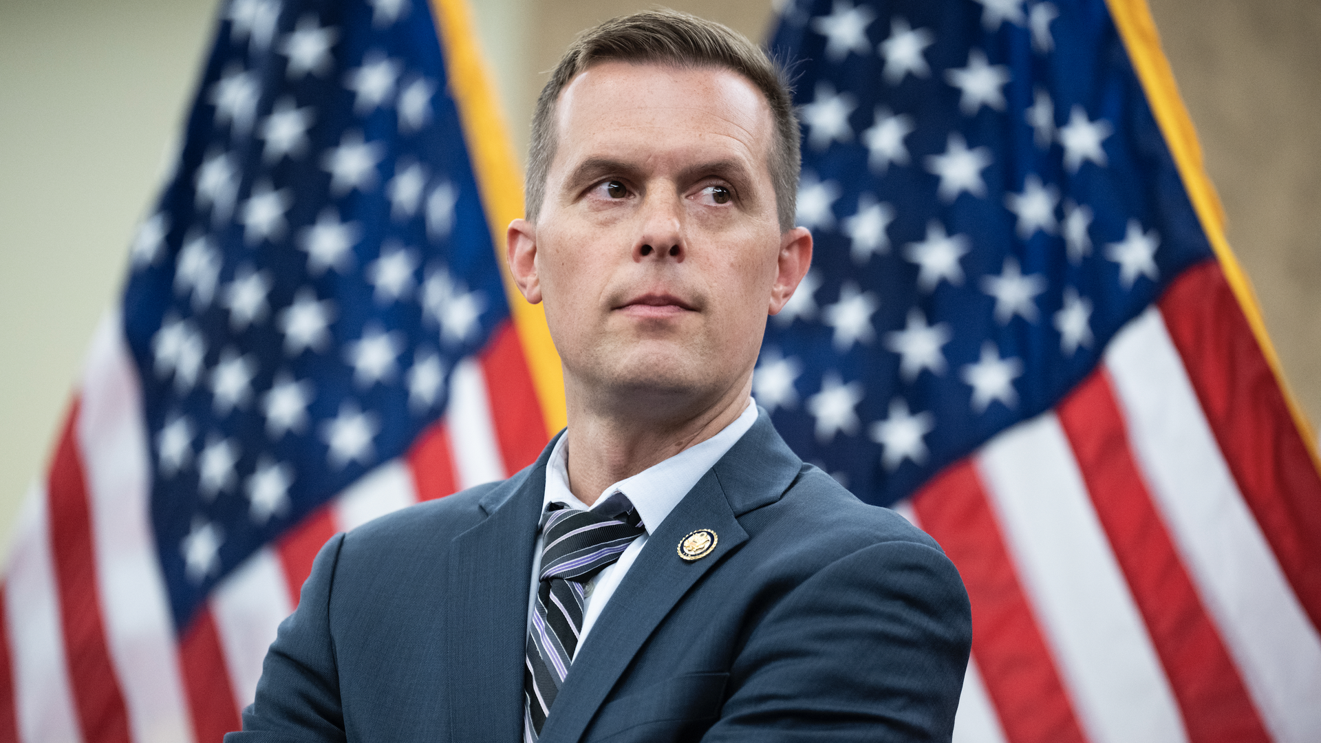 A man in a blue suit and striped tie with a lapel pin, arms crossed, stands in front of large American flags, looking to the left.