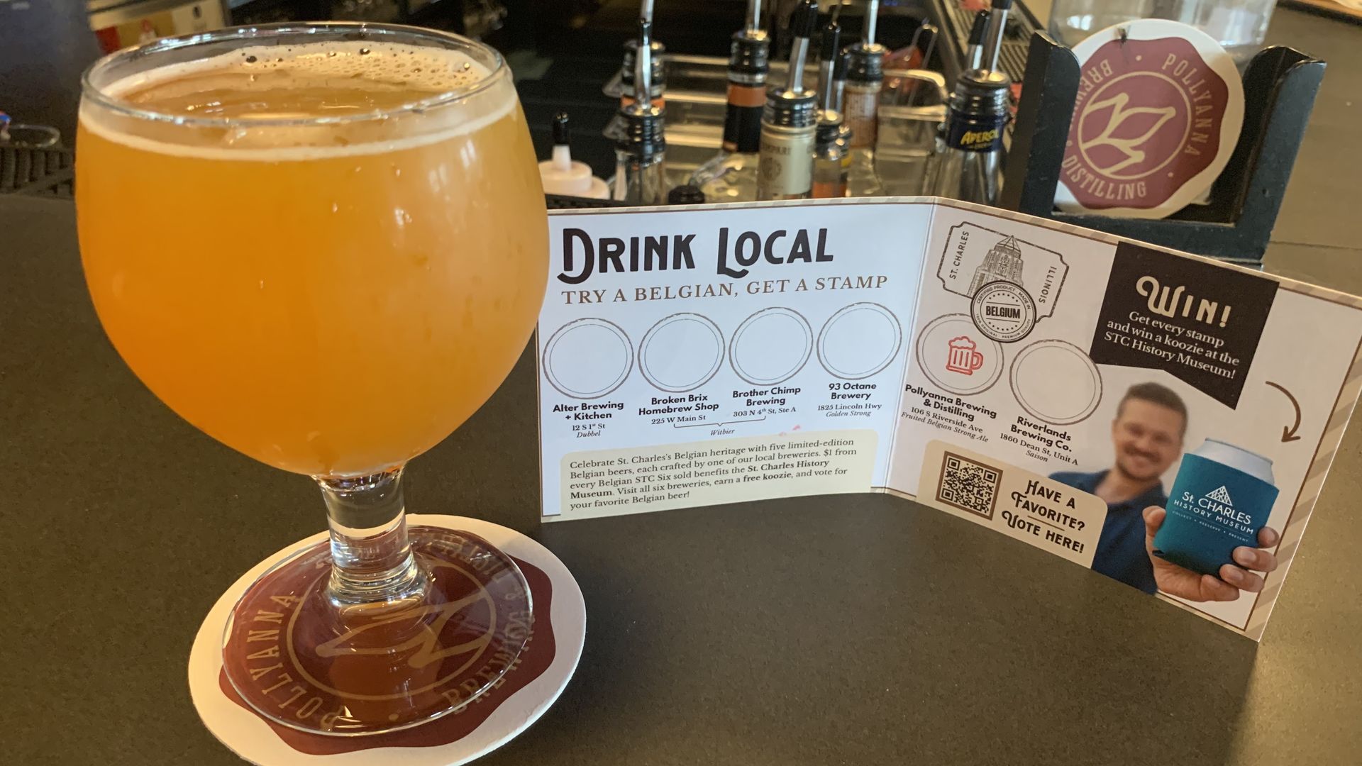 Foreground shows a hazy amber beer in a stemmed glass on a coaster; bar counter with beer taps and bottles in the background, and a Drink Local promotional flyer on the counter.