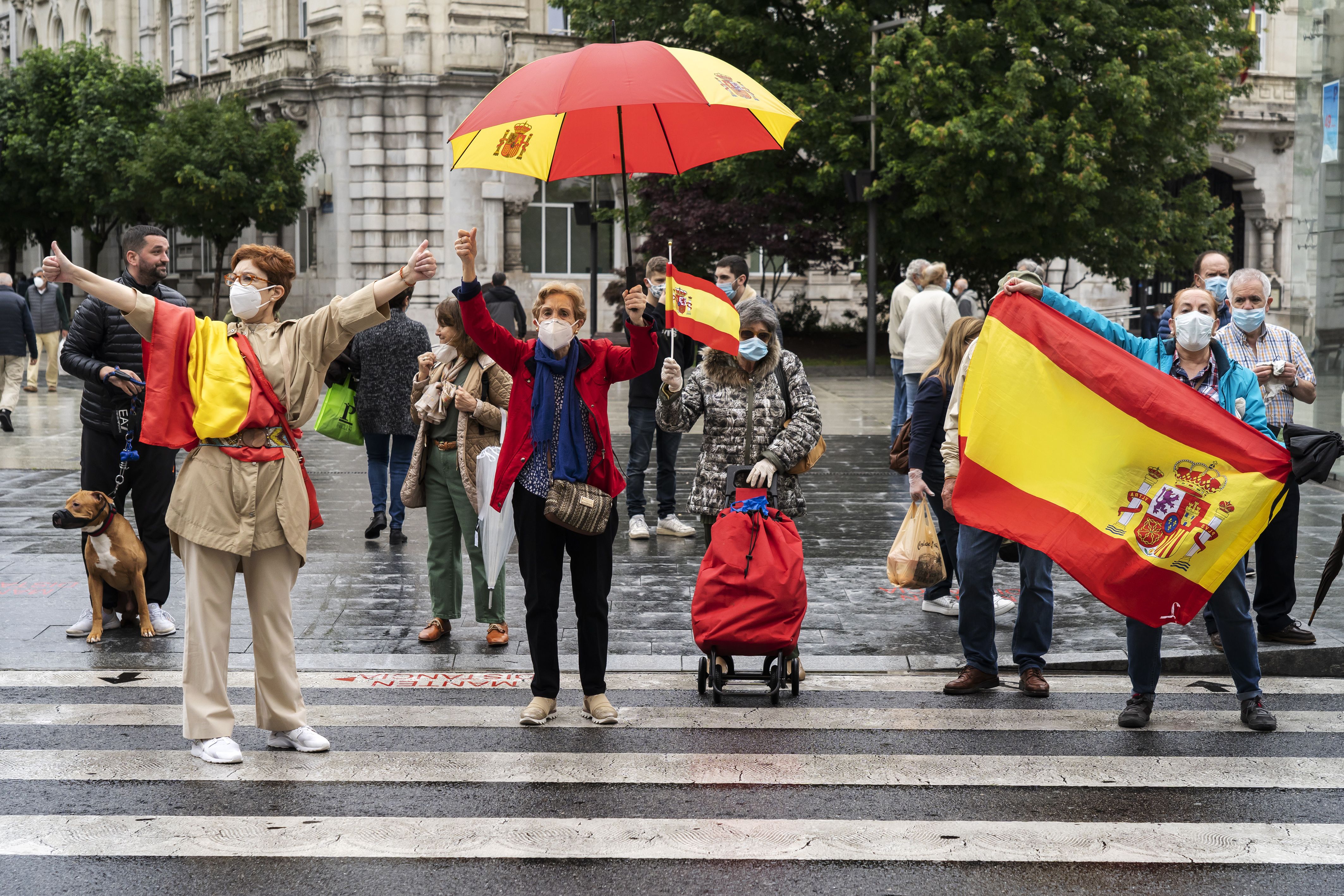 In this image, a group of people holding flags and umbrellas decorated with Spain's flag stand in a crosswalk