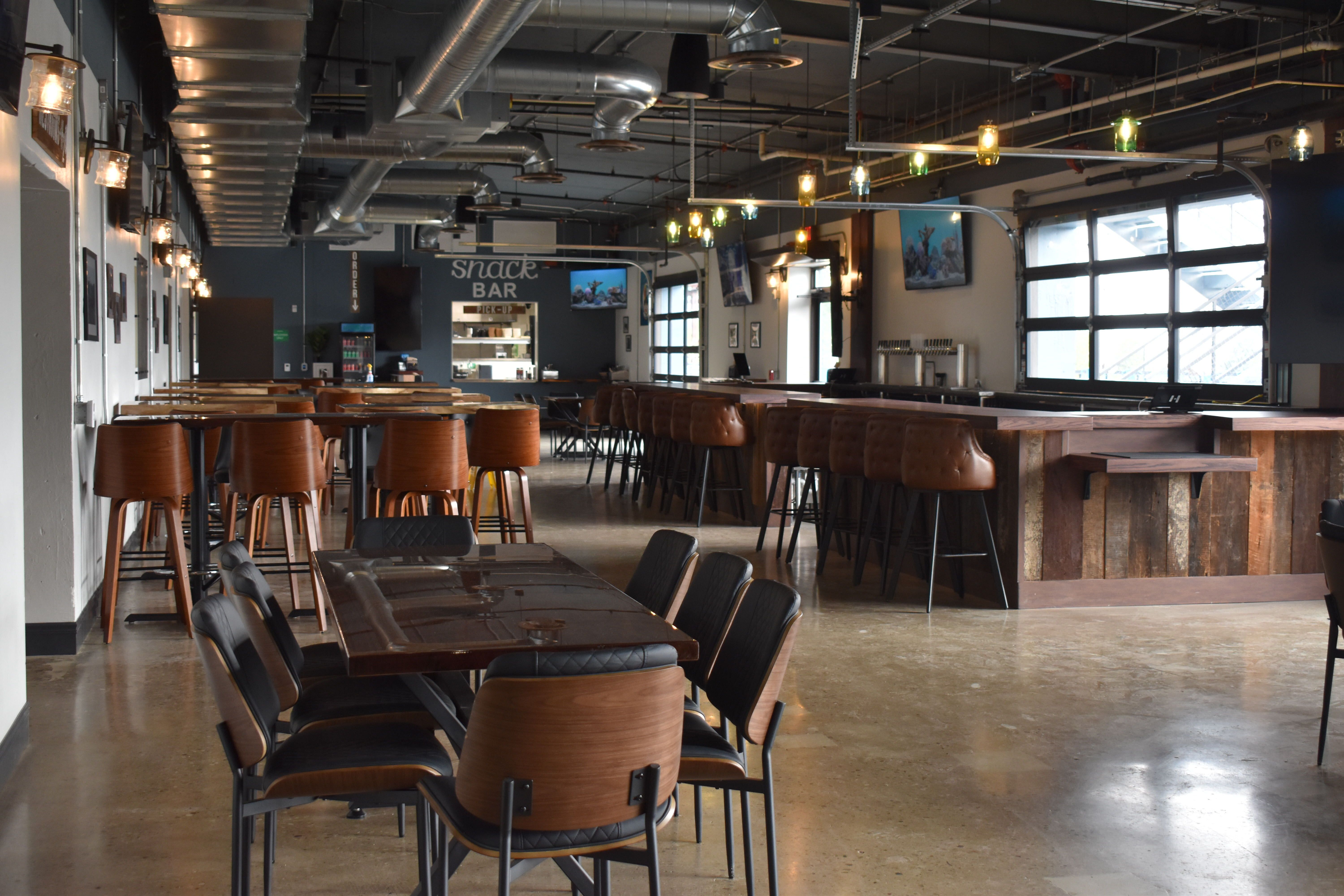 Empty modern bar and snack area with wooden tables, brown leather and black chairs, industrial ceiling with ducts, and windows letting natural light inside.