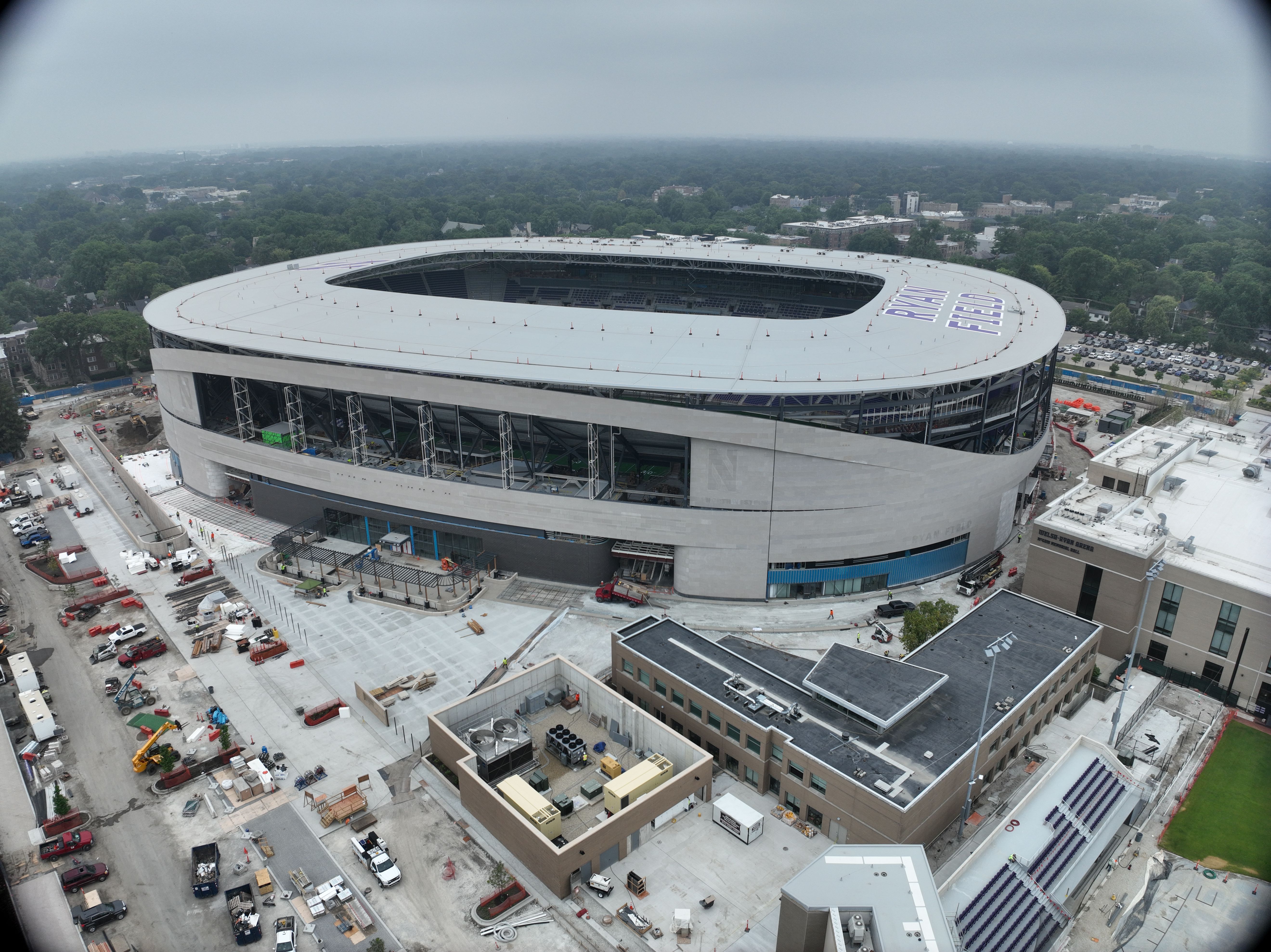 An aerial view of a large circular stadium under construction, with a white ring roof and open center, surrounding scaffolding, construction equipment, and a green tree-filled landscape in the distance.