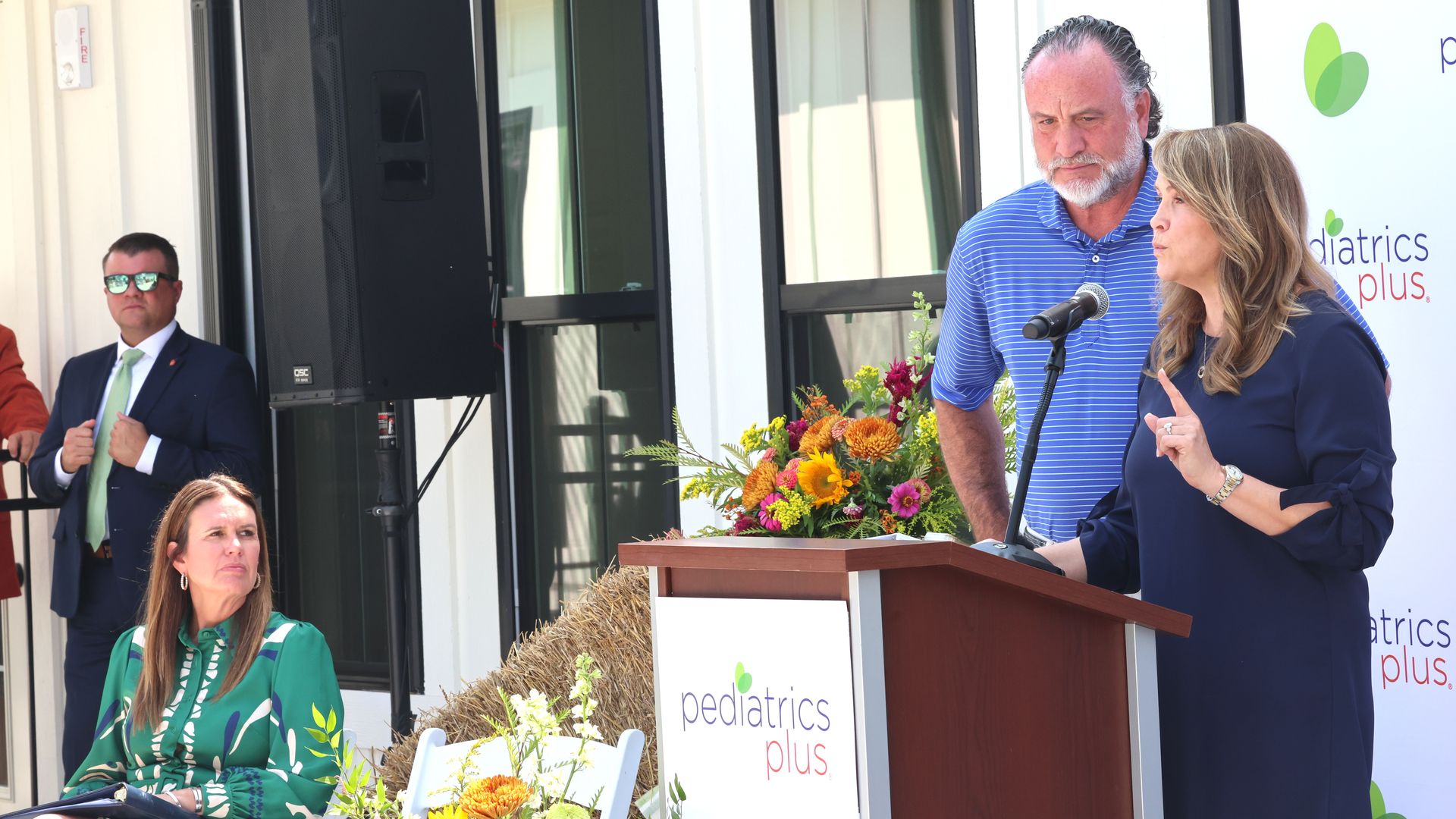 Woman in navy dress speaking at podium with "pediatrics plus" logo, man in blue striped shirt beside her, woman in green patterned dress sitting, man in suit with green tie stands nearby.