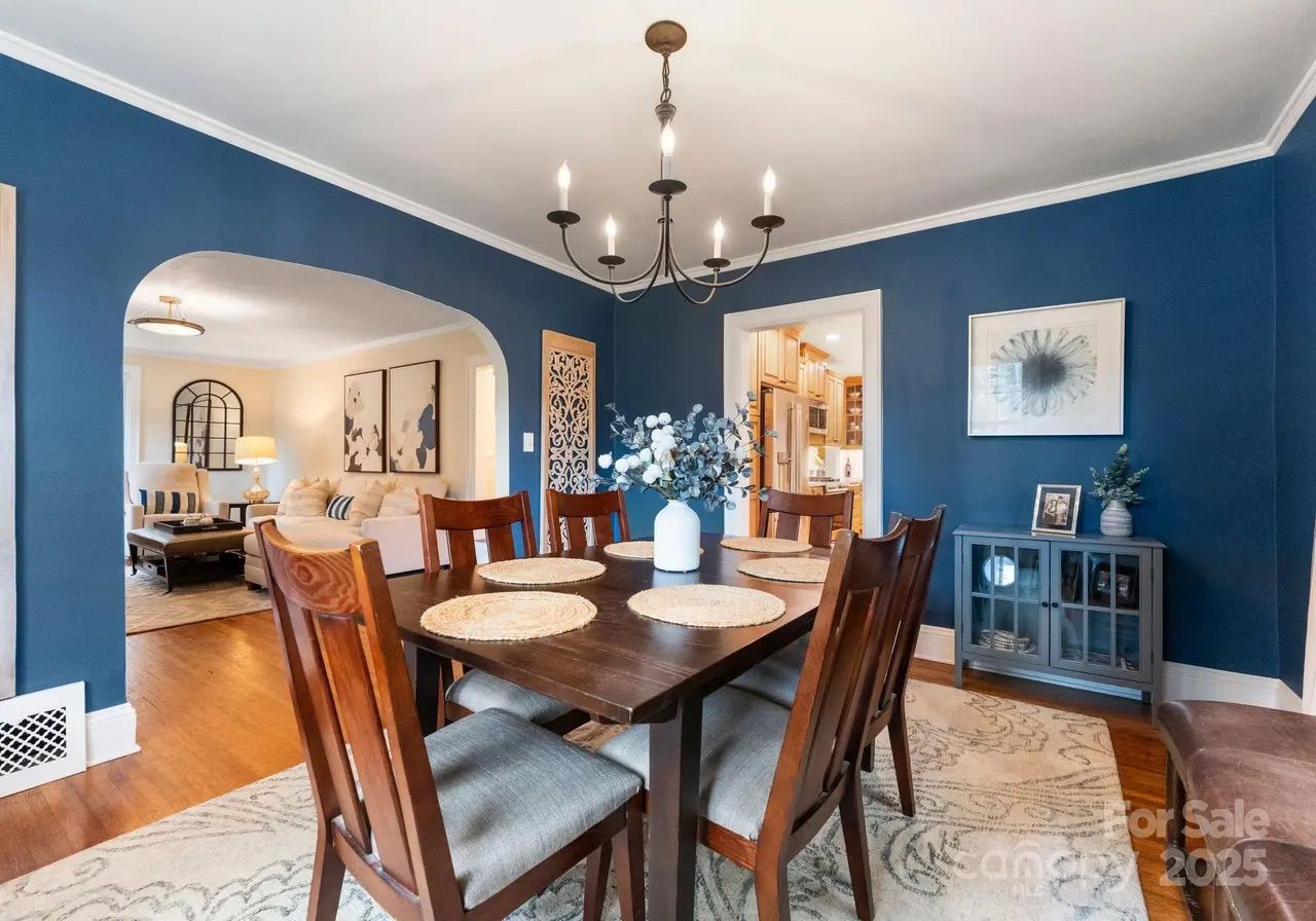 Dining room with blue walls, wooden table set with six chairs and round placemats, white vase with flowers, chandelier overhead, adjacent living room with beige sofa and wall art.
