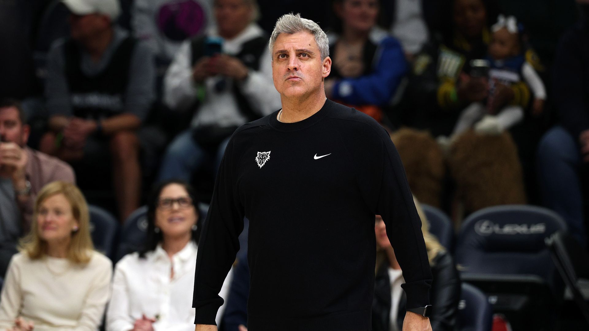 A man with short gray hair and a serious expression stands in a black athletic shirt with a white Nike swoosh; a blurred crowd sits in the stands behind him.