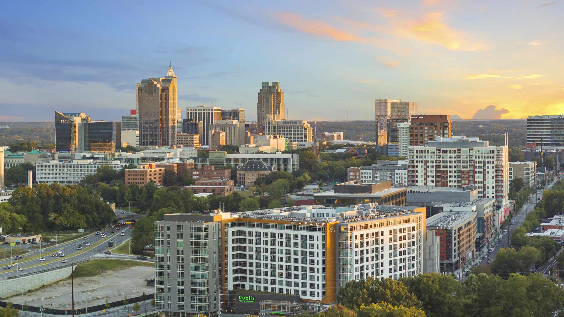 Sunset city skyline with tall glass towers, mid-rise buildings, and green trees. A curved road and a Publix store are visible in the foreground beneath a warm orange sky.