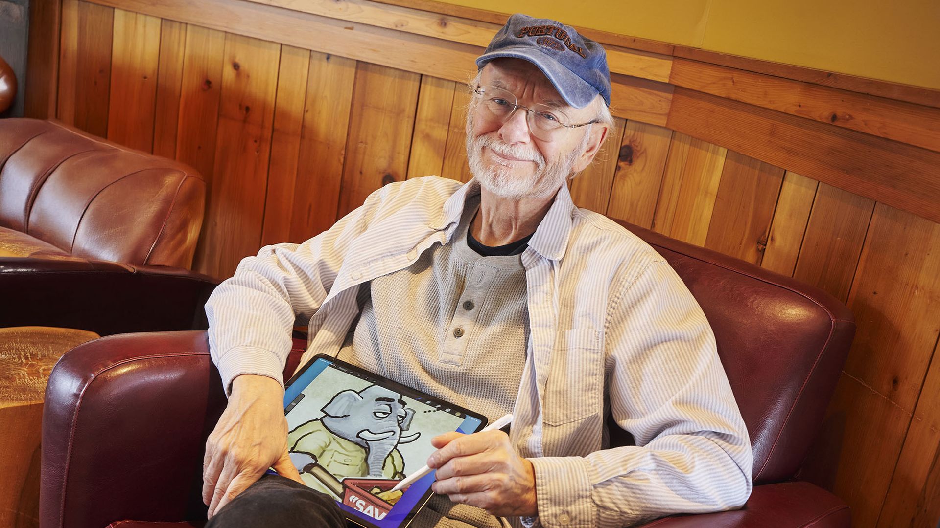 Older man with gray beard and glasses sits on a red leather couch in a wood-paneled room, wearing a blue cap and striped shirt, holding a tablet with a cartoon image.